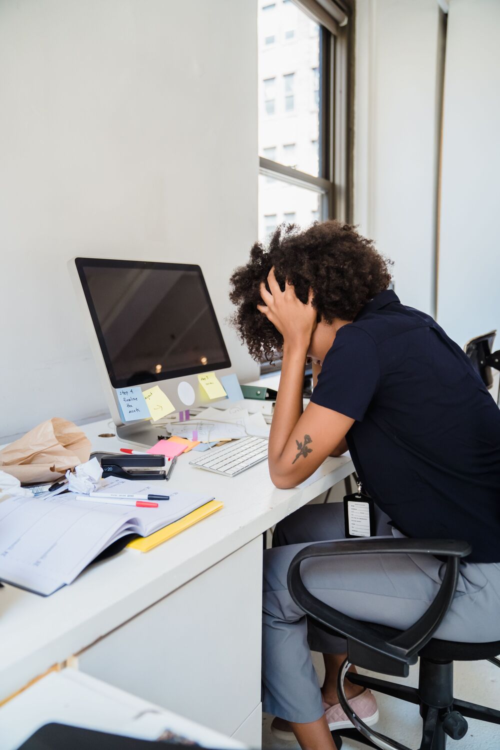 Woman with curly hair sitting at a cluttered desk, holding her head in stress