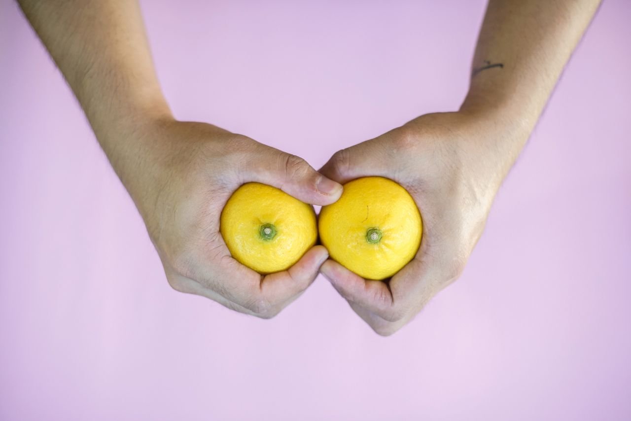 Two hands holding a pair of lemons against a pink background, used to symbolise breasts