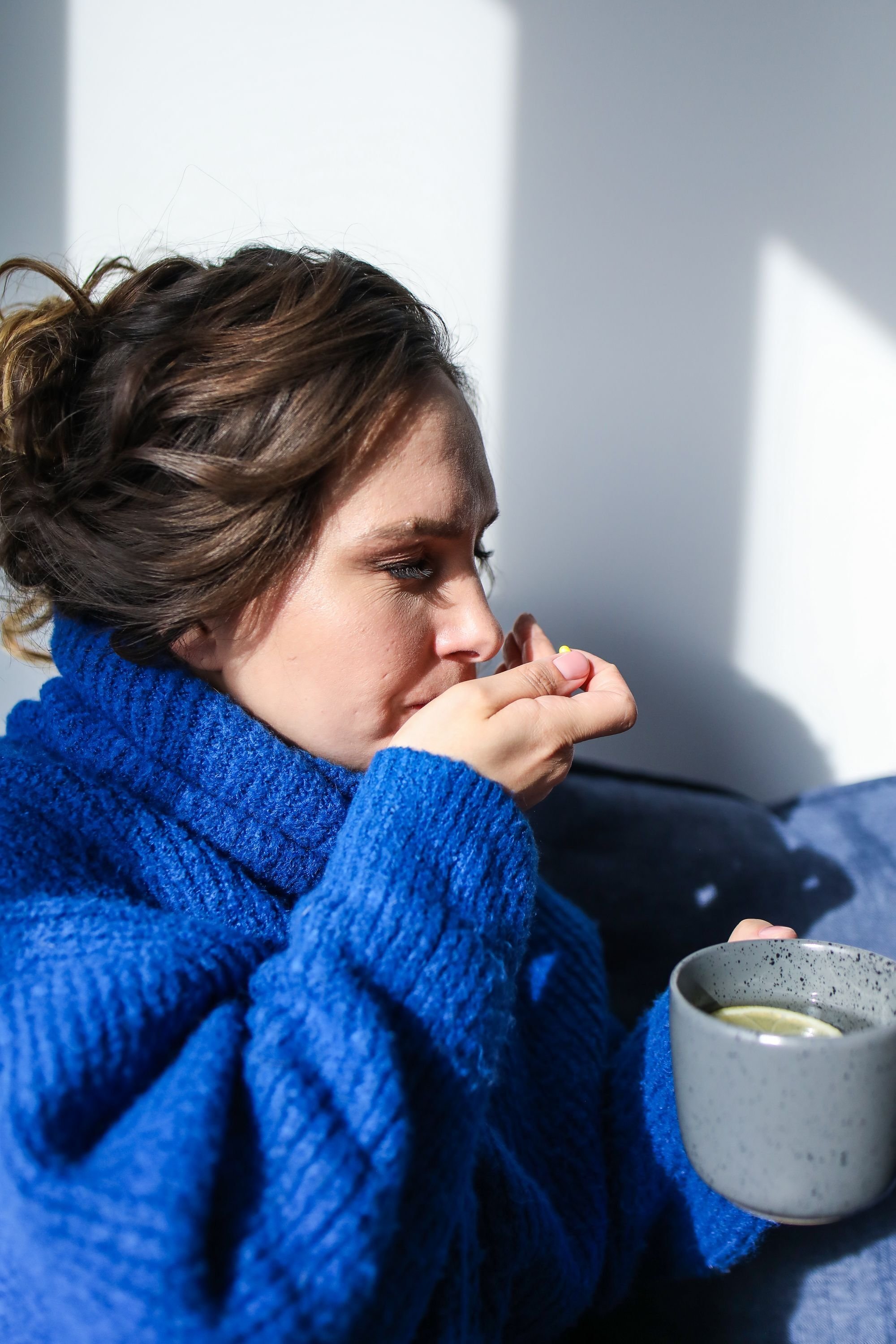 Woman in a blue jumper taking a pill while sitting on a sofa