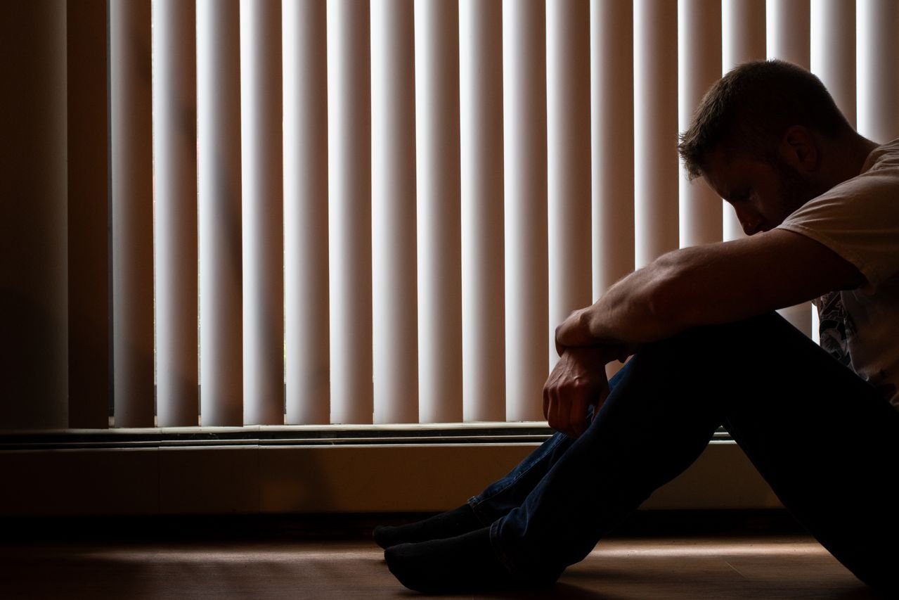 Silhouette of a man sitting on the floor by a window, head down, appearing distressed