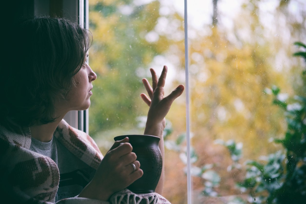 Fall melancholy mood concept. Wistful woman sitting near window and looking at autumn landscape outside in rainy day