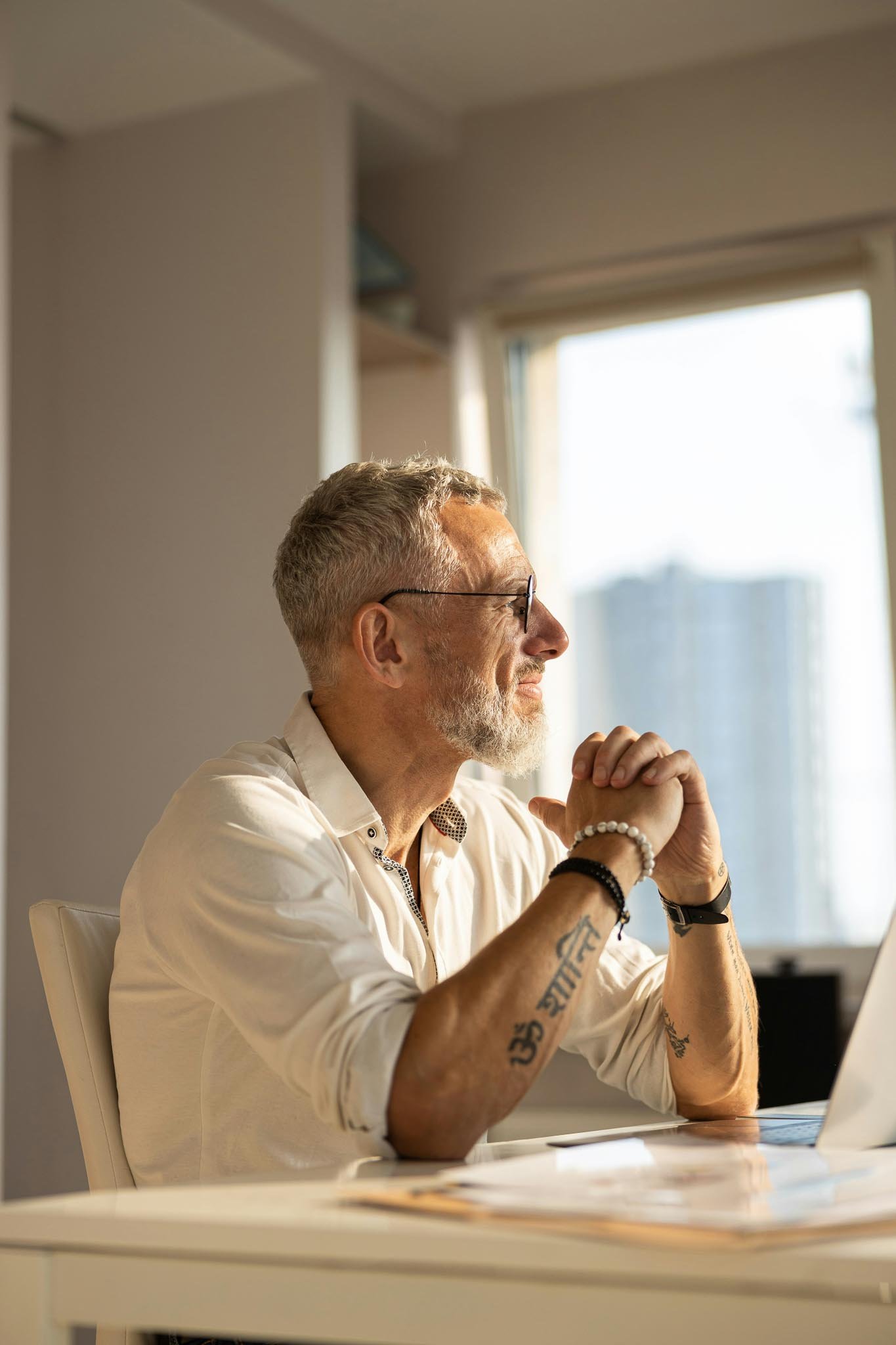 Grey-haired man with glasses and tattoos smiling thoughtfully while looking out a window with hands clasped