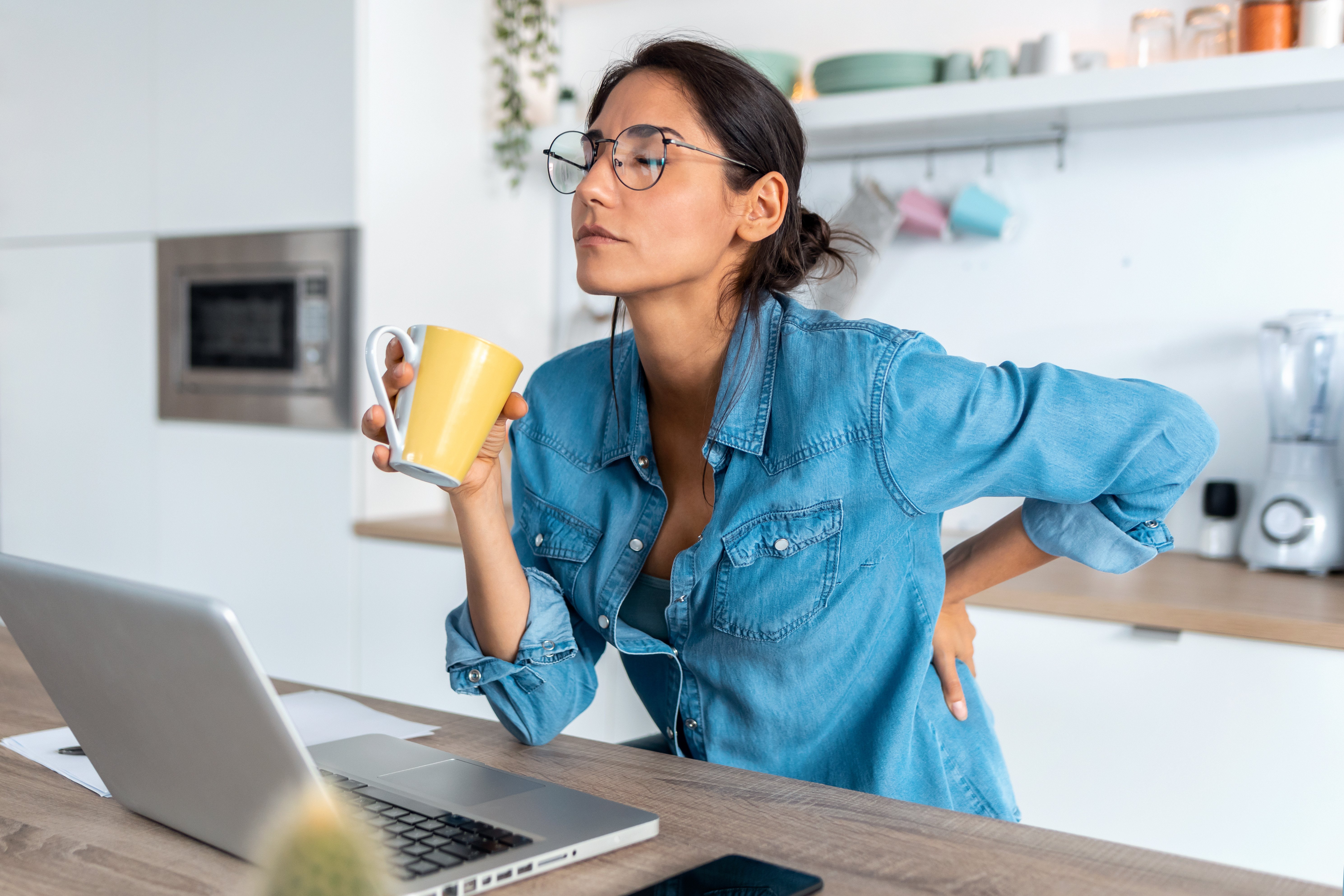 Woman working from home in her kitchen, holding a mug with a hand on her lower back as if experiencing back pain