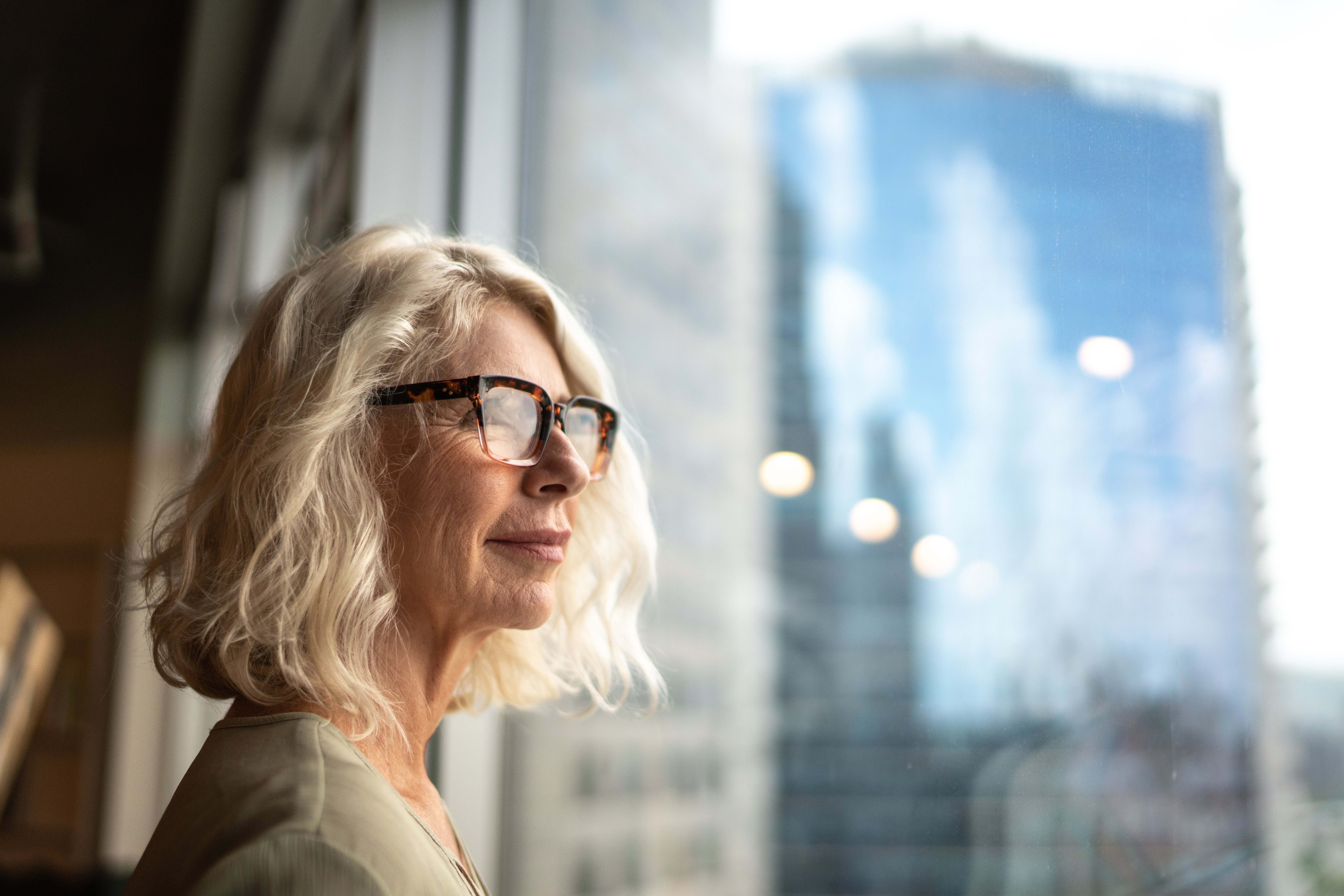 Older woman with grey hair and glasses looking out a window at city high-rises