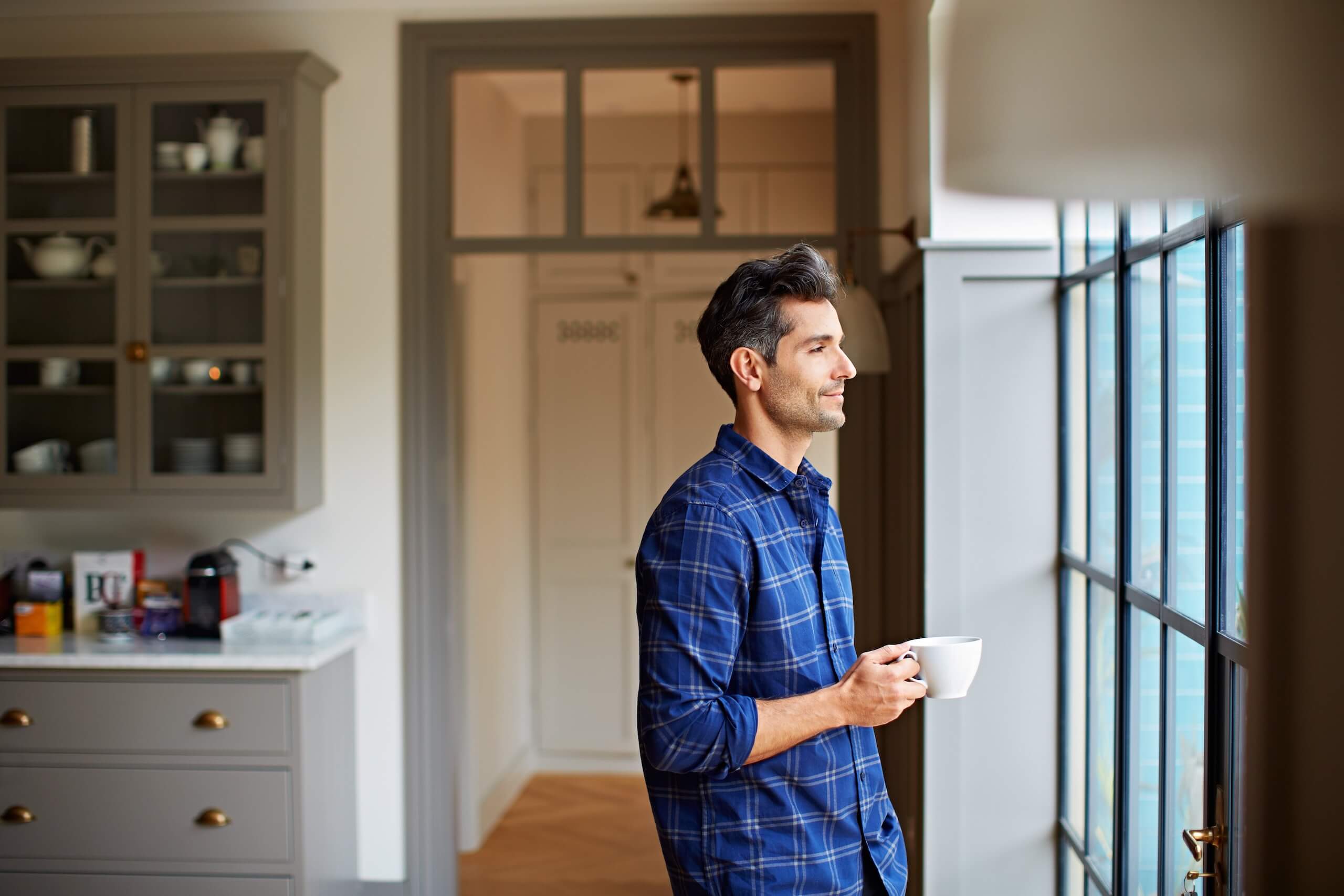 Man in a checked shirt holding a mug and looking out of a window