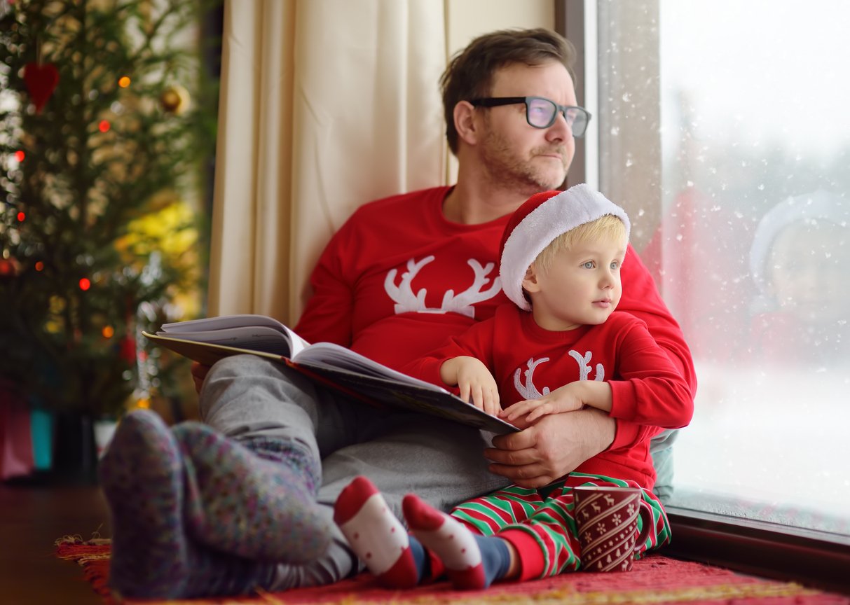 Little boy wearing Santa hat and his father are reading the book, drinking hot cocoa and looking out the window on snowfall during Christmas time