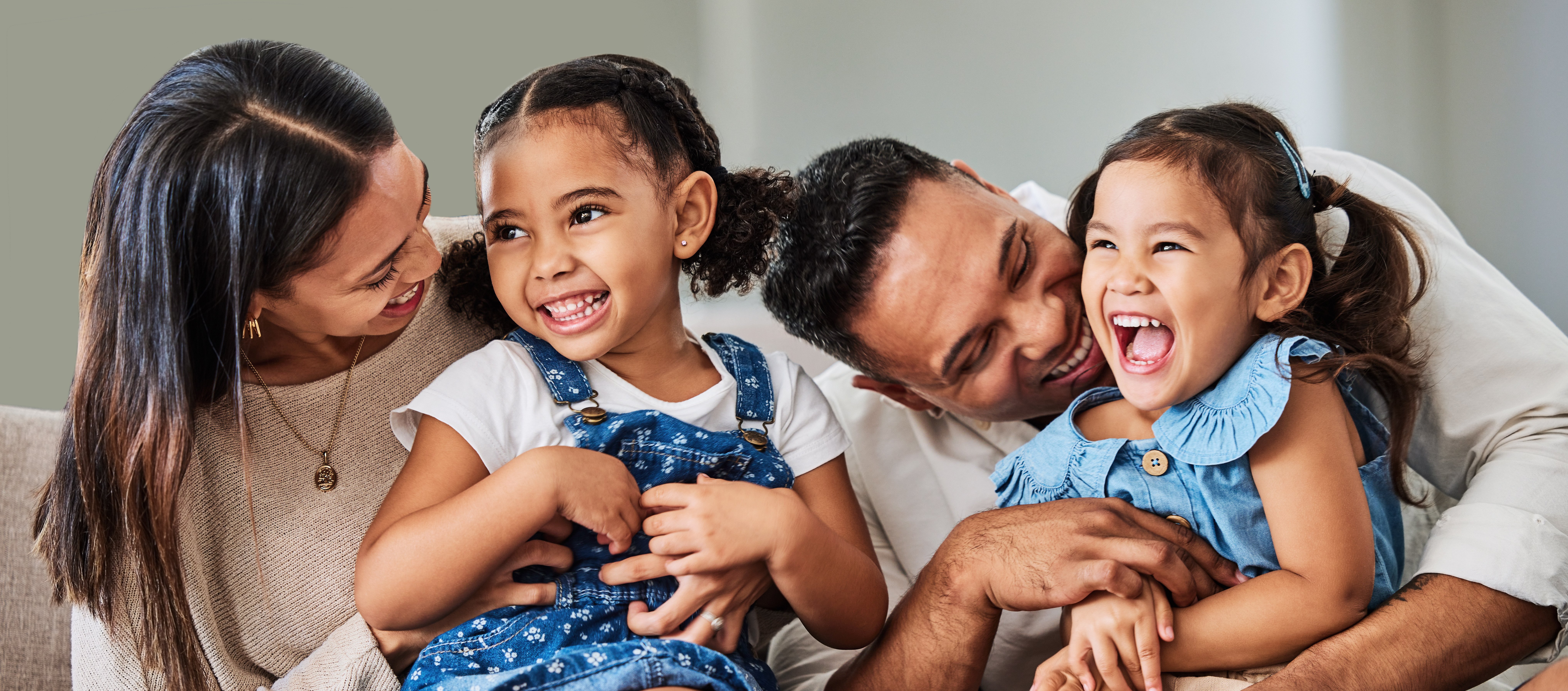 A family laughing together on a sofa, with two parents and two young daughters wearing matching denim outfits.