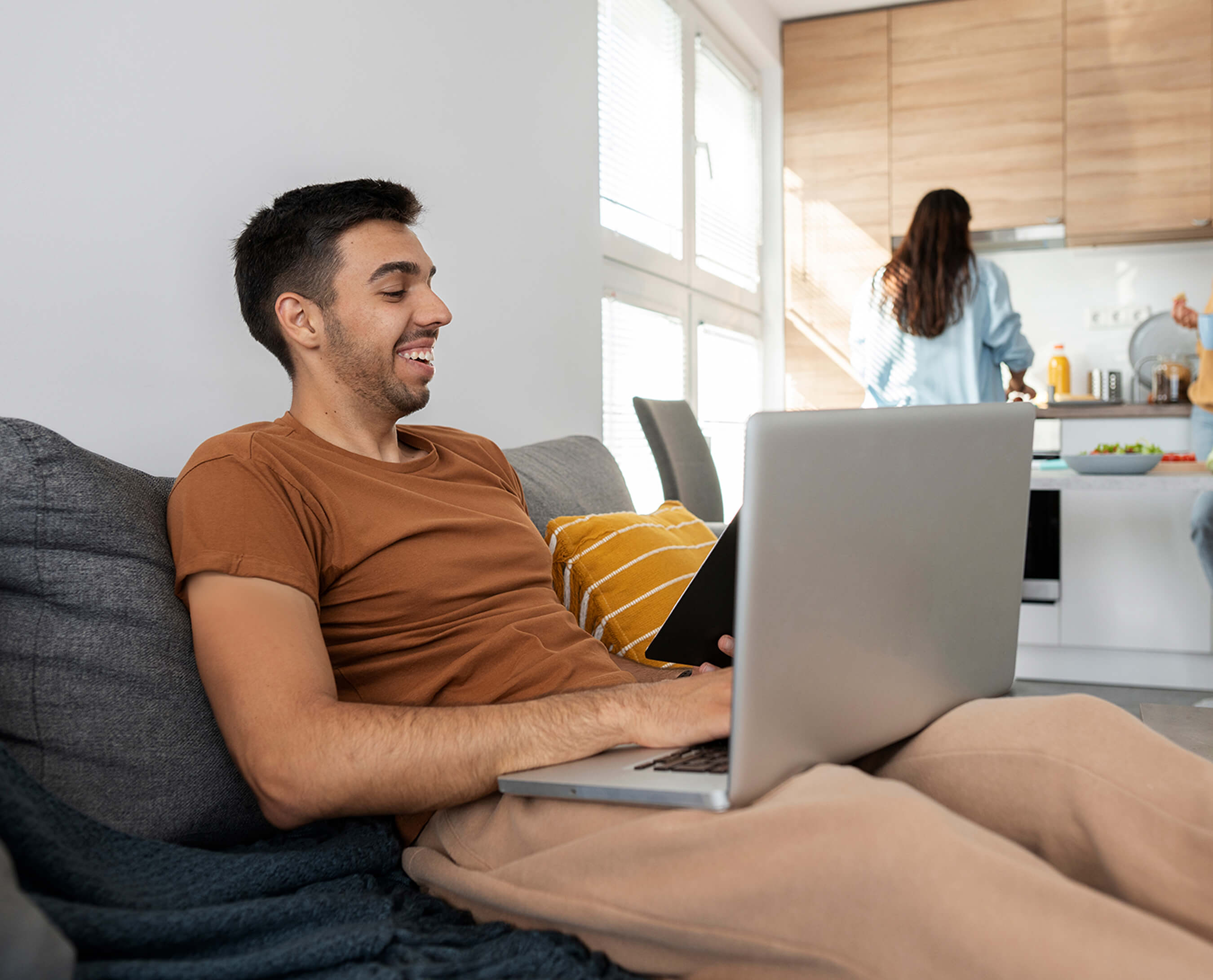 smiling man on a video call with his laptop, with a person in the kitchen behind him