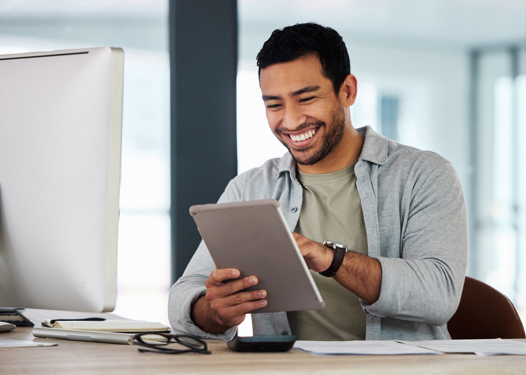 Smiling man using a tablet at his desk in front of a computer monitor