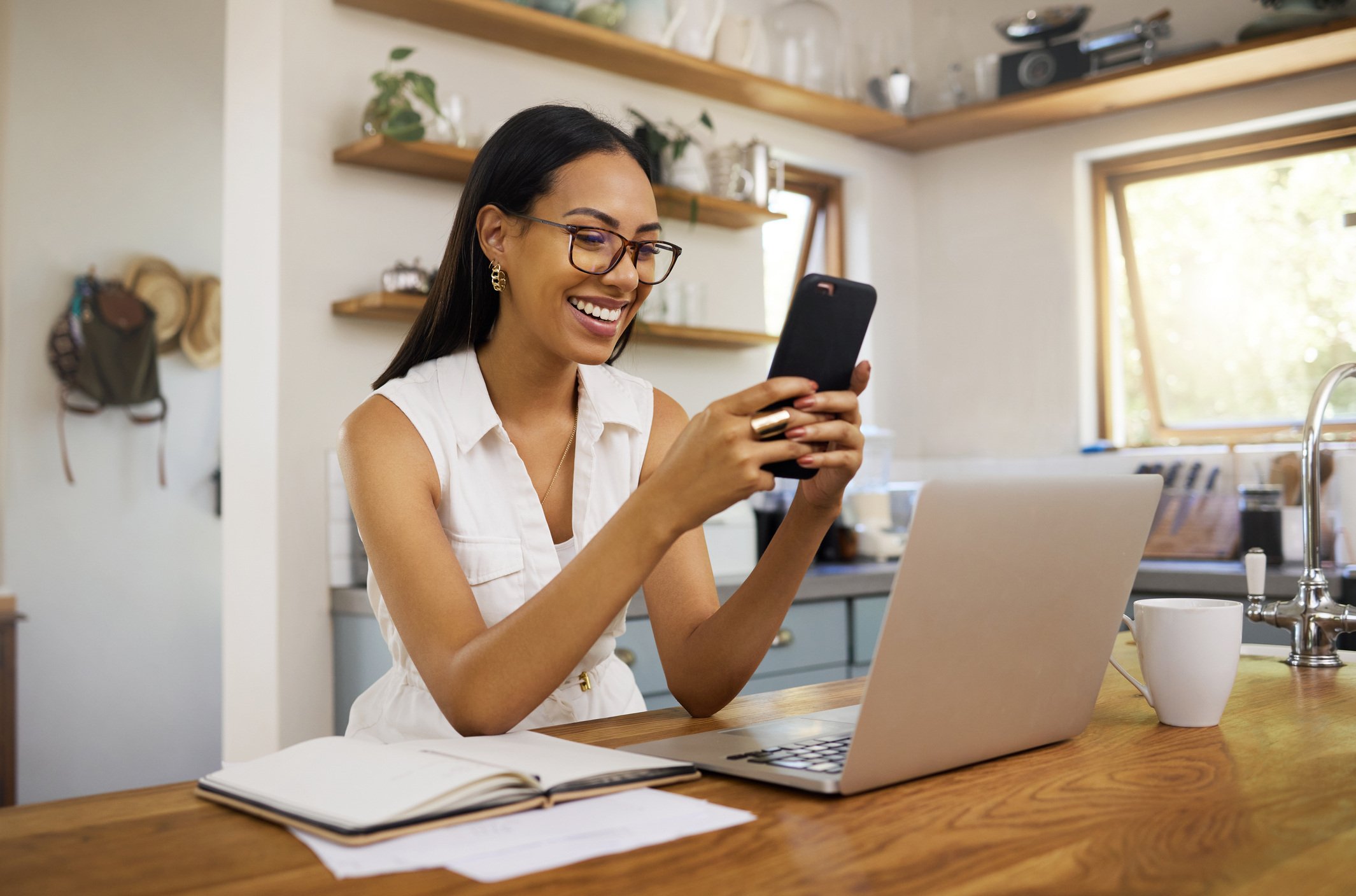 Smiling woman checking her phone at a laptop in a kitchen