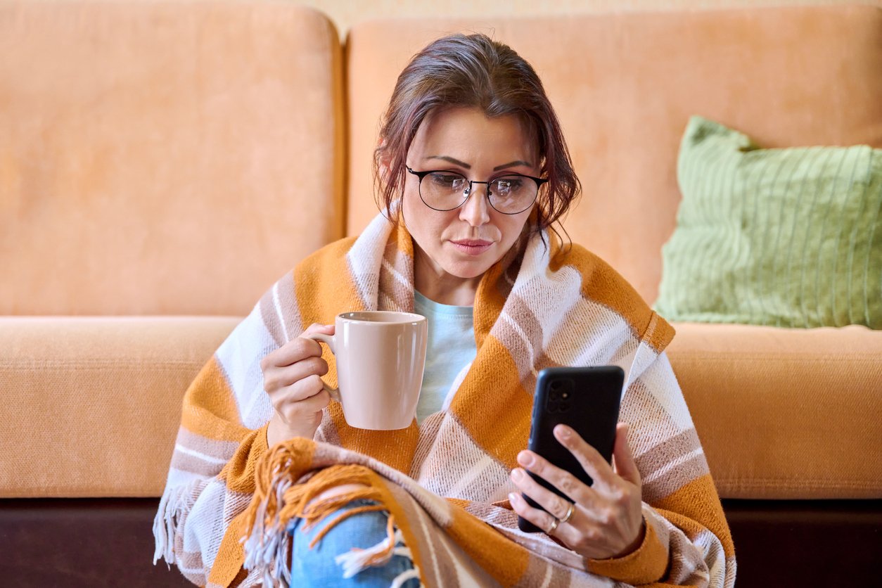 Woman under a woolen warm blanket with a cup of hot tea using a smartphone