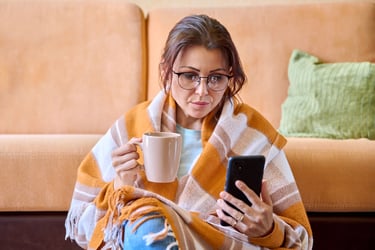 Woman under a woolen warm blanket with a cup of hot tea using a smartphone