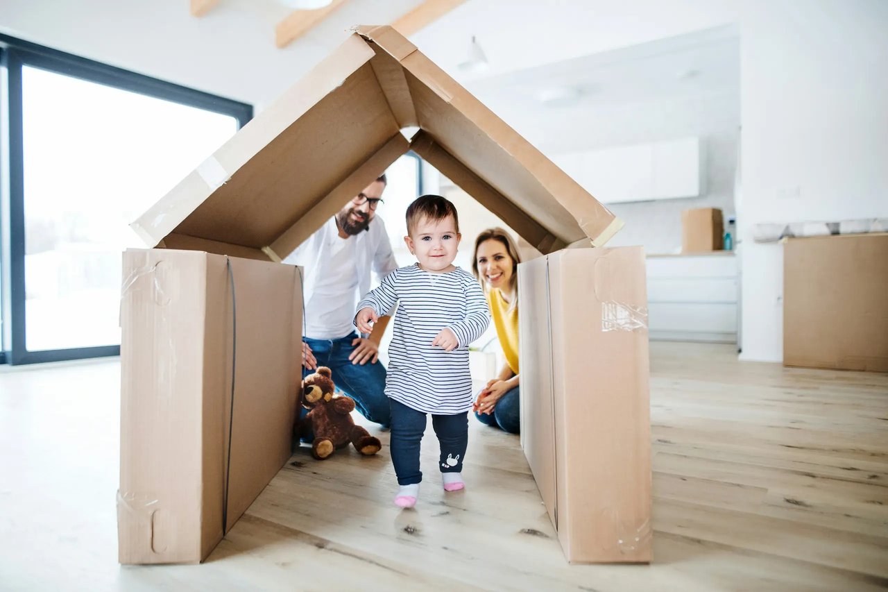 Two adults and a child standing inside a large cardboard house frame in a bright empty room