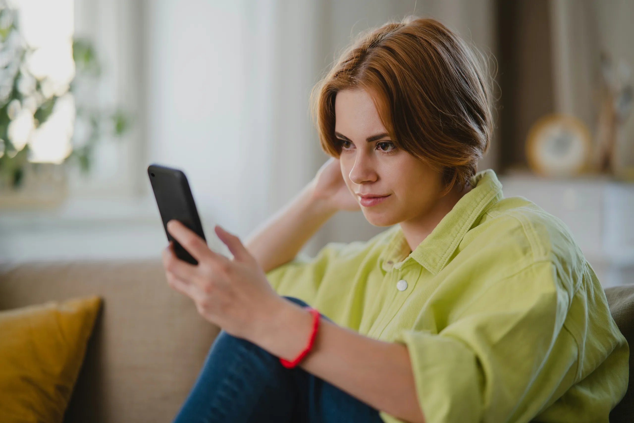 Woman with a bob haircut looking thoughtfully at her phone on a sofa