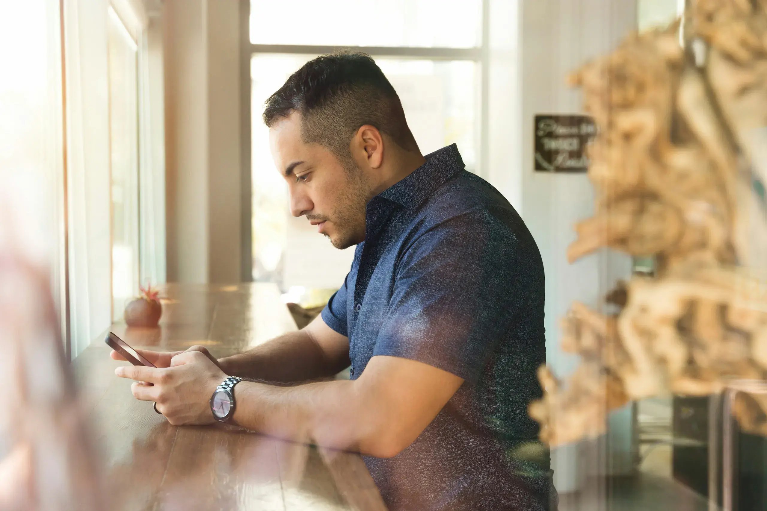 Man checking his phone while seated at a wooden counter by a window