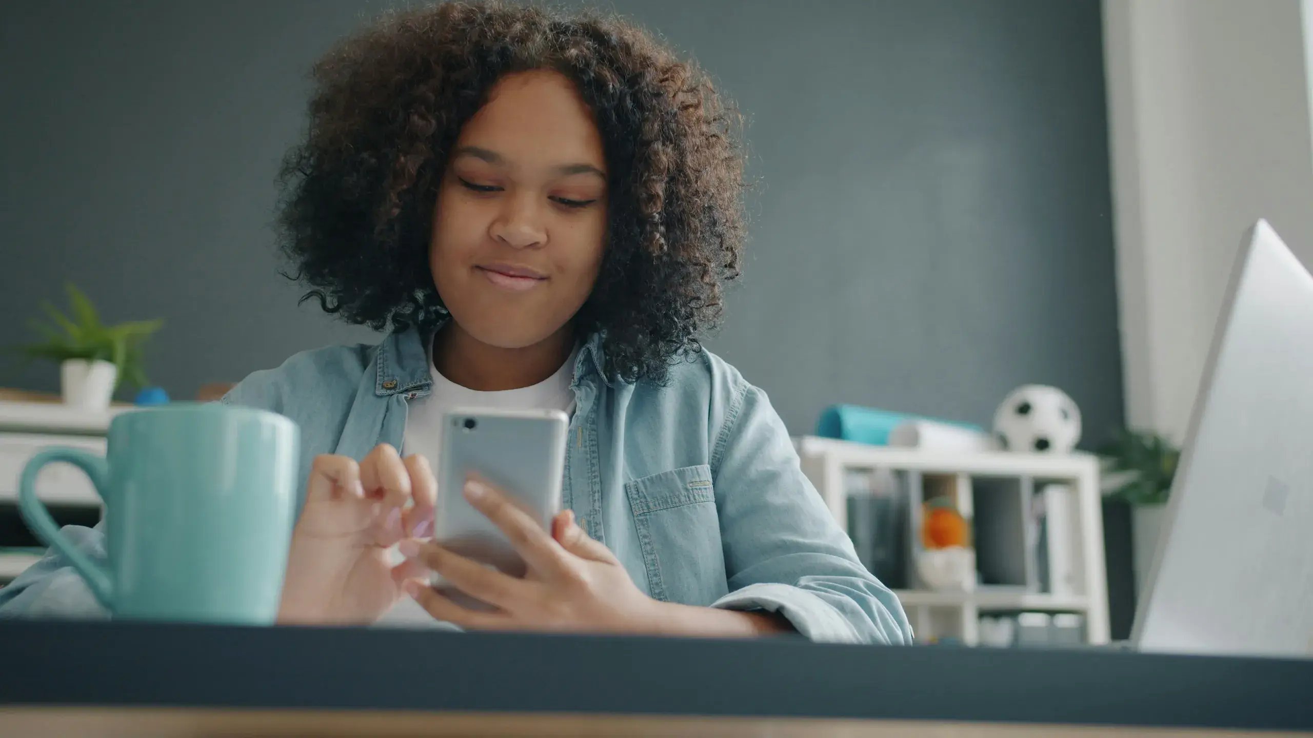 Smiling teenage girl with curly hair checking her phone at a desk with a mug