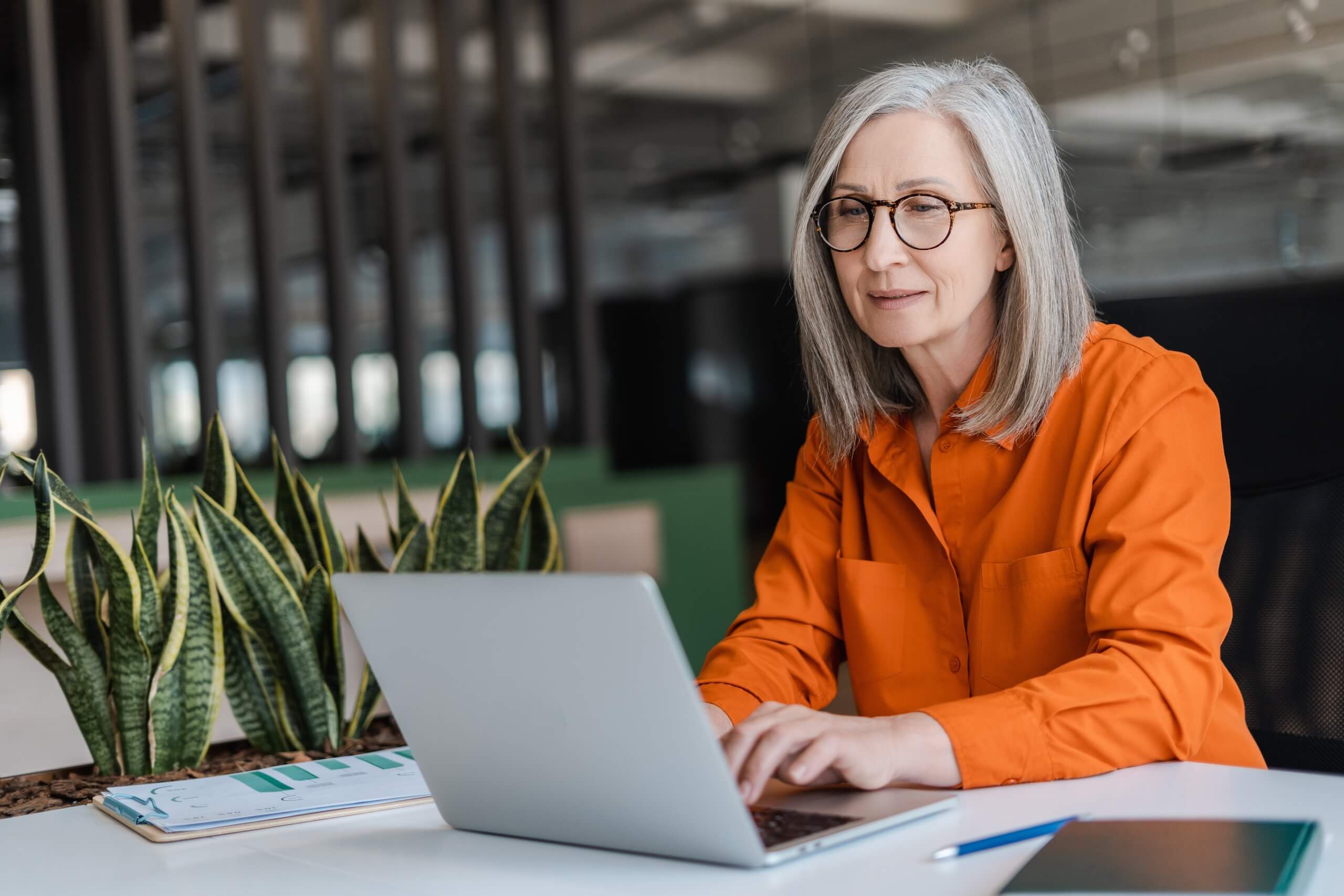 Older woman with grey hair and glasses working on a laptop at a desk surrounded by office plants