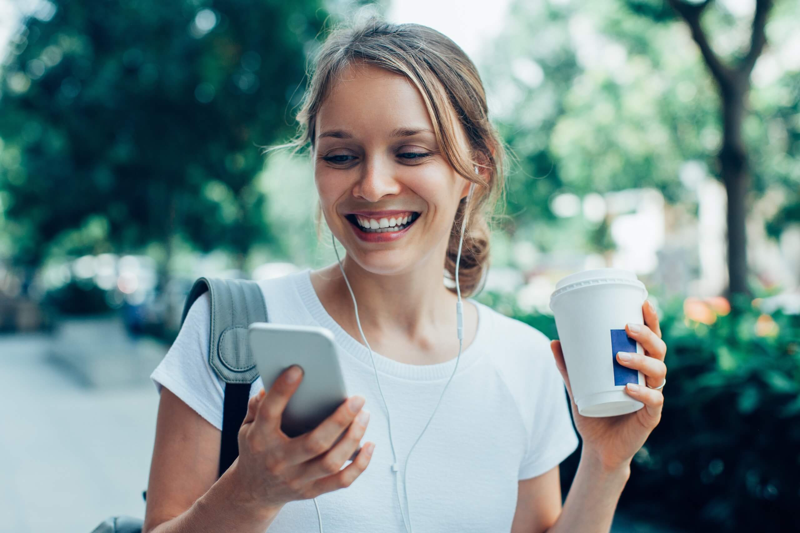 smiling woman outdoors holding a coffee cup and looking at her phone