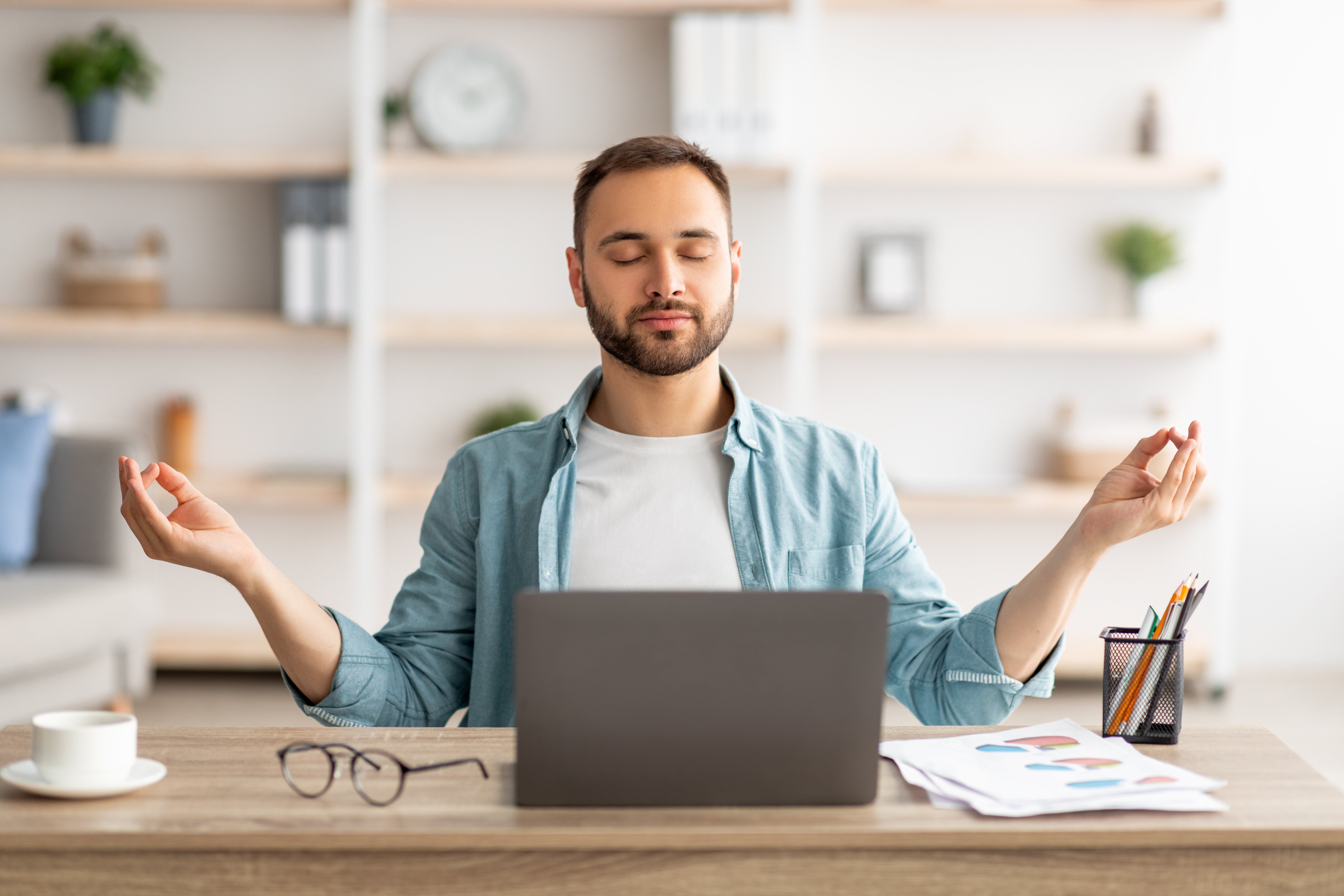 Workplace stress management. Calm Caucasian man meditating with closed eyes in front of laptop pc at home office