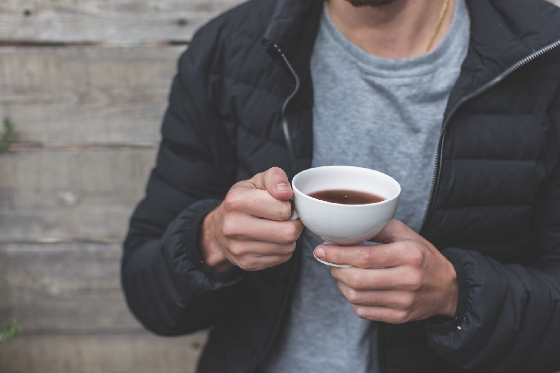 A person in a black puffer jacket and grey t-shirt holds a white cup of tea in both hands against a wooden background