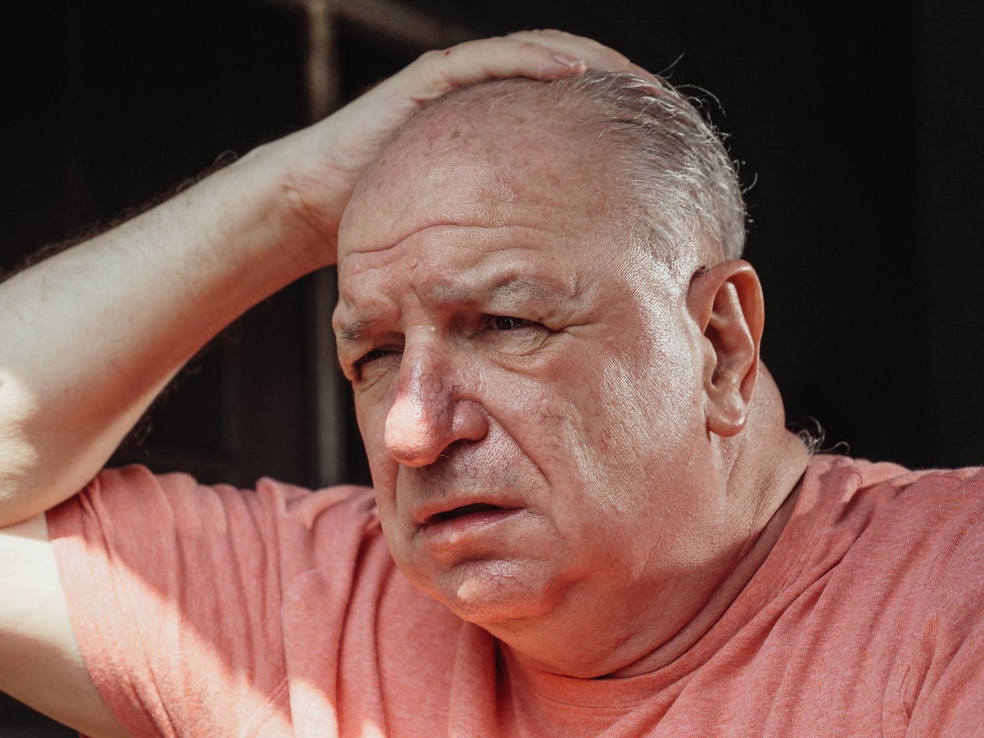 An older bald man outdoors rests his hand on his head, squinting with a concerned or confused expression.