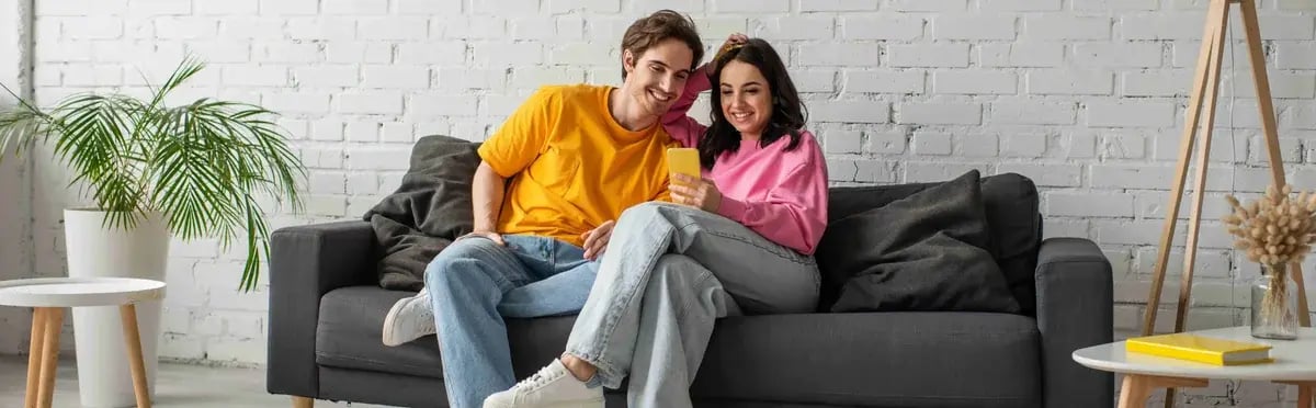positive young couple sitting on couch and looking at cell phone in living room, banner