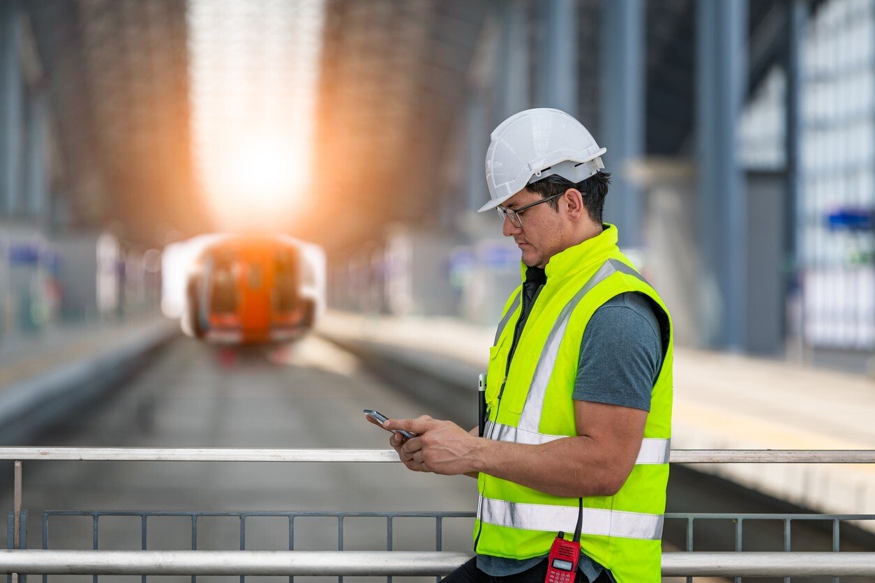 Railway worker in a hi-vis vest and hard hat checking a tablet on a train platform