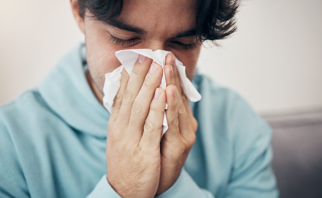 A man in a light blue hoodie blows his nose into a tissue with his eyes closed, appearing to have a cold or allergies