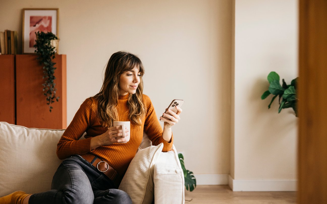 Woman sitting on a sofa at home holding a mug and checking her phone