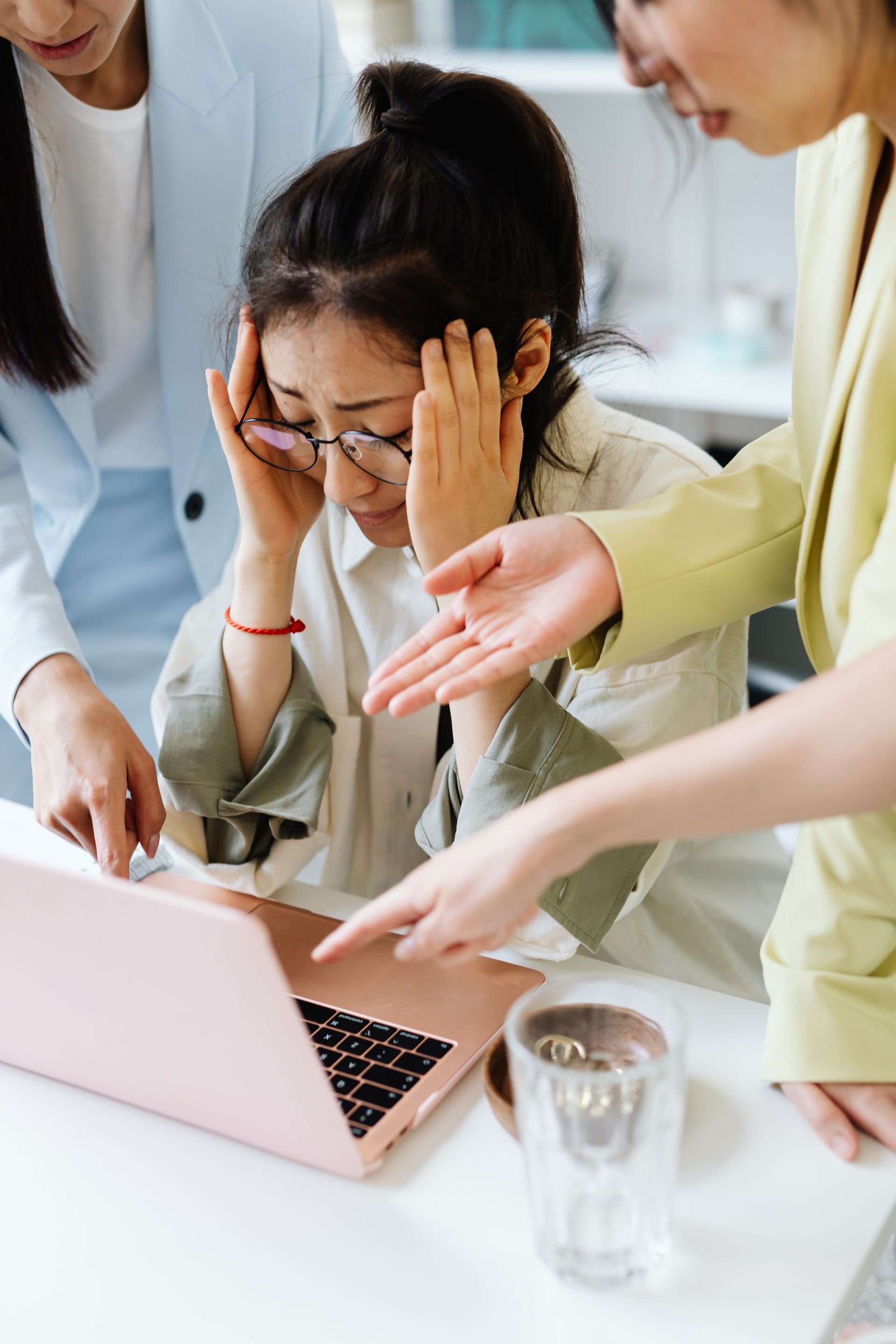 Overwhelmed woman holding her head at a laptop while two colleagues gesture and point around her