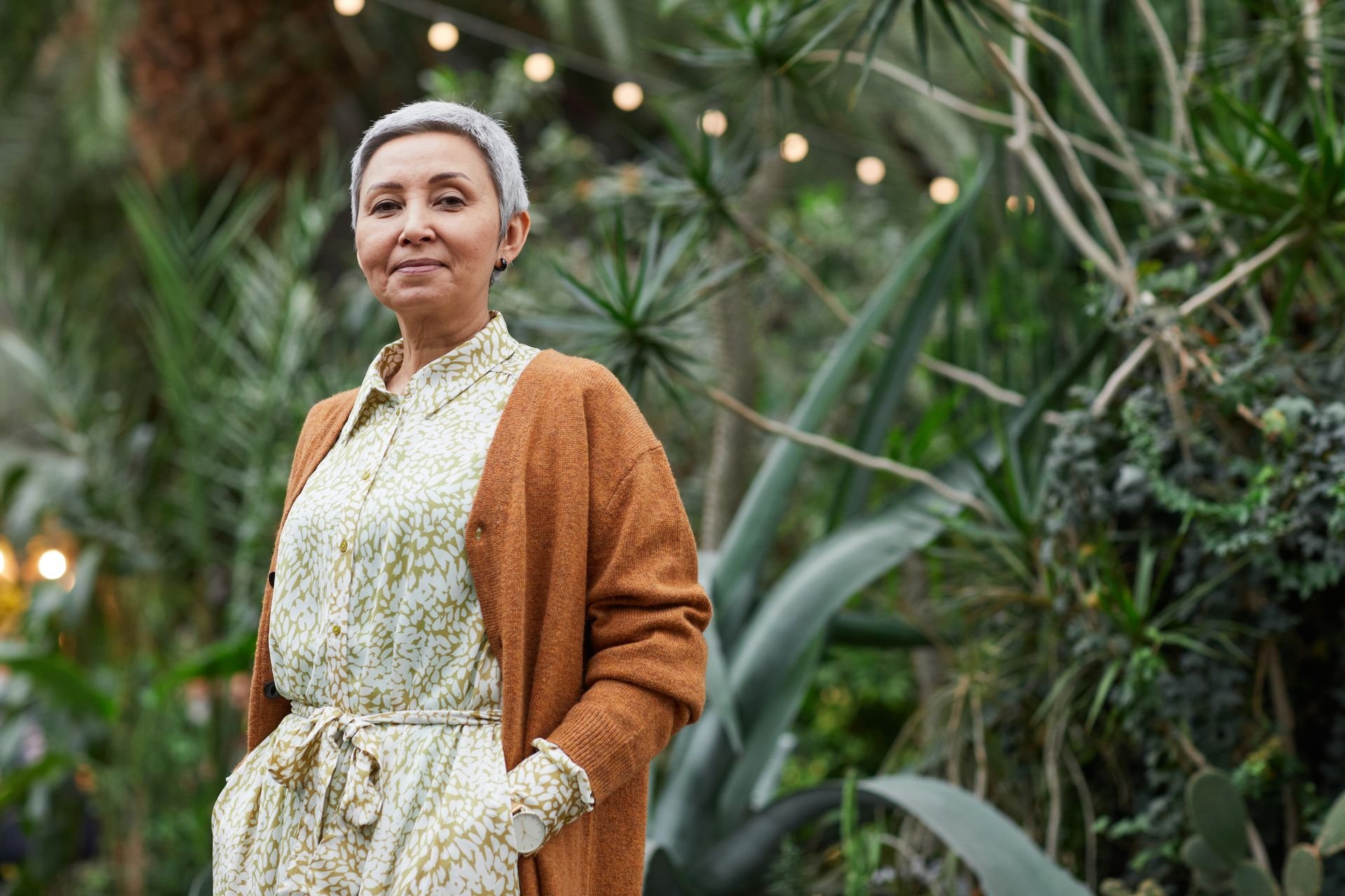 Smiling older woman with grey hair wearing a cardigan, standing among green foliage