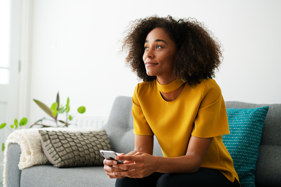 A smiling woman with curly hair, wearing a mustard yellow top, sits on a couch holding a phone and looking up and to the side.