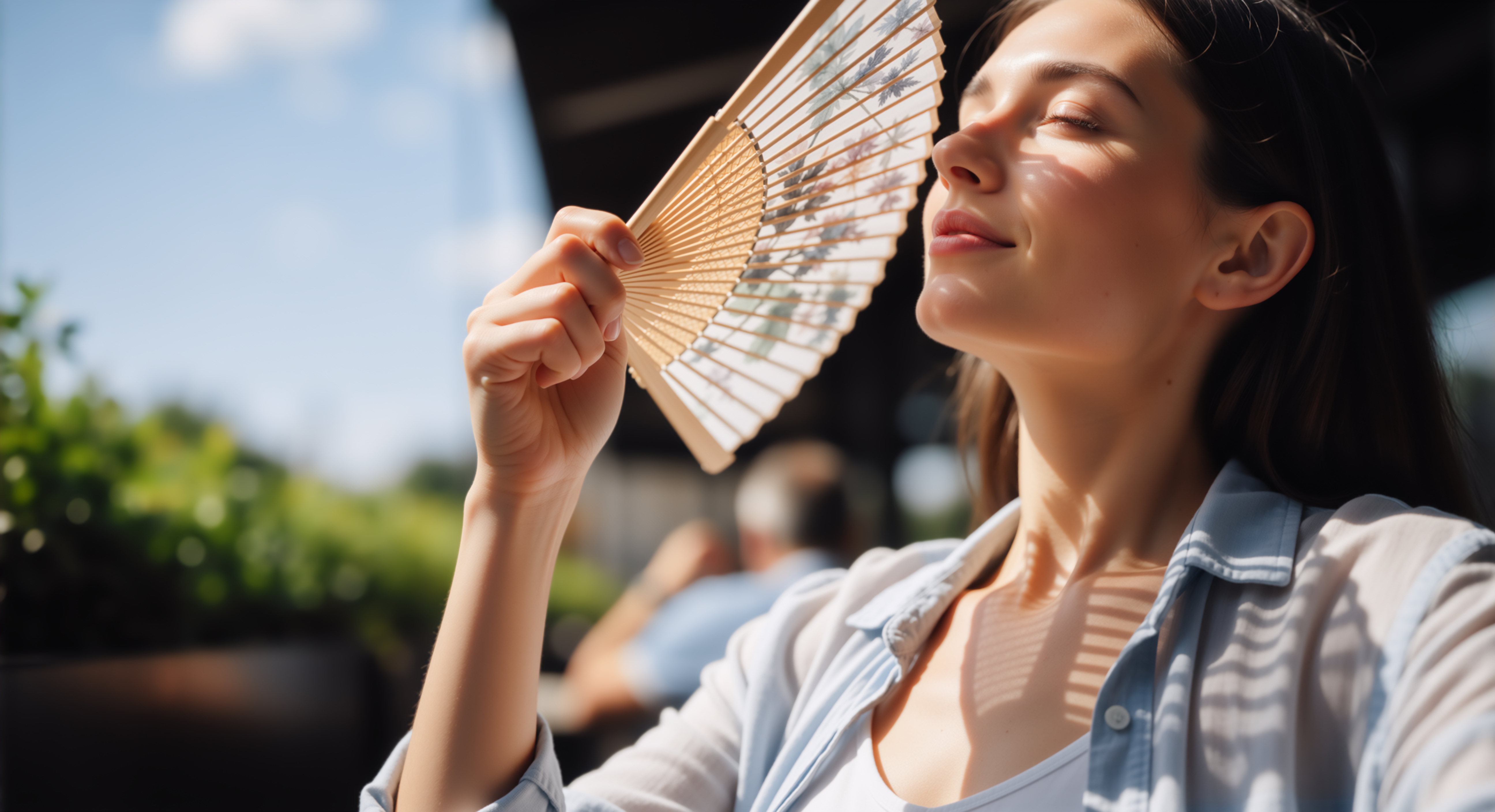 A woman outdoors fanning her face with a hand fan, appearing to cool herself, with her neck and shoulders visible in close-up.