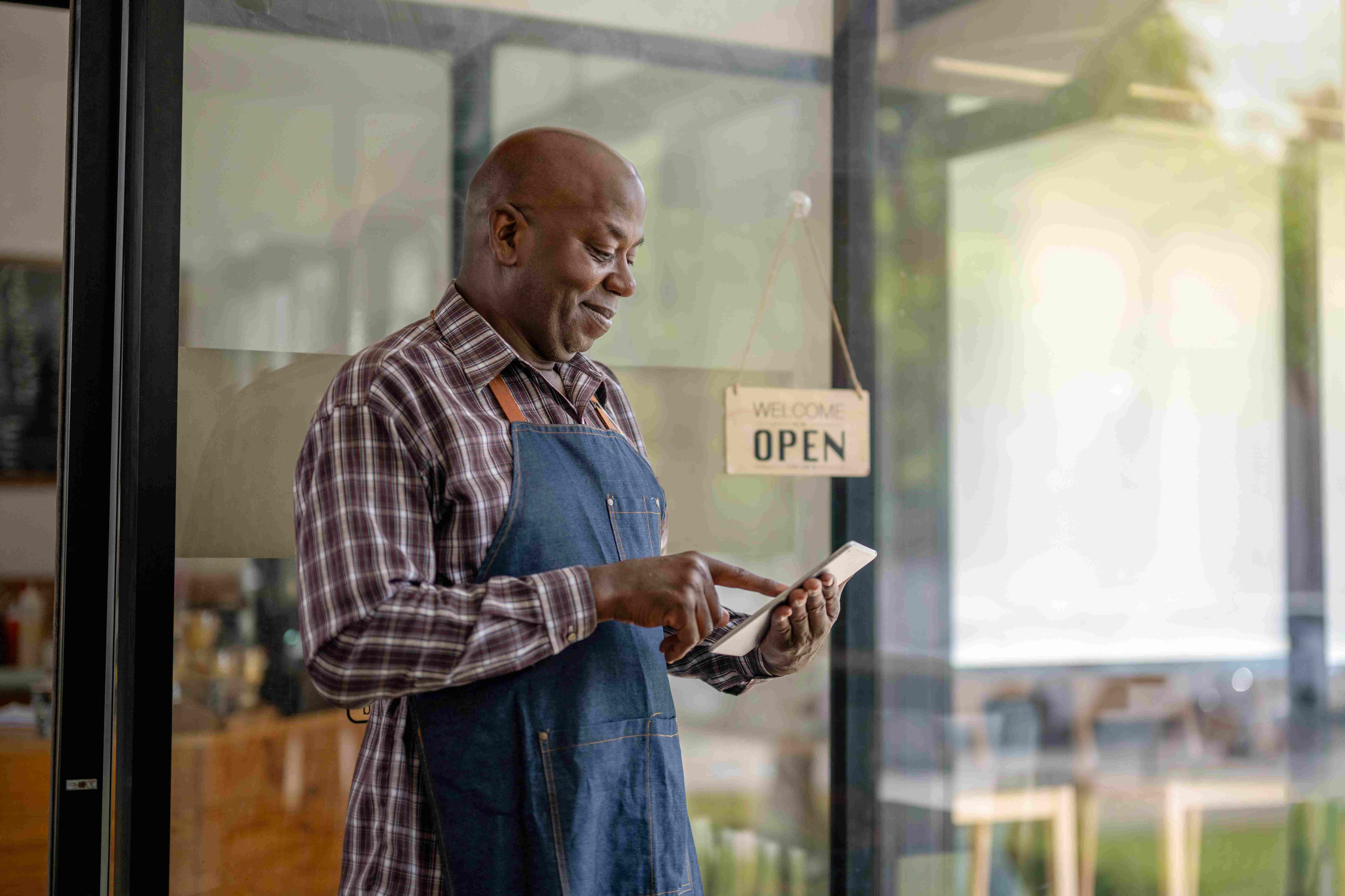 A smiling man wearing an apron stands near a shop's glass door with an 'Open' sign, looking at a tablet or phone in his hands. An orange 'Featured' badge is in the top right corner.