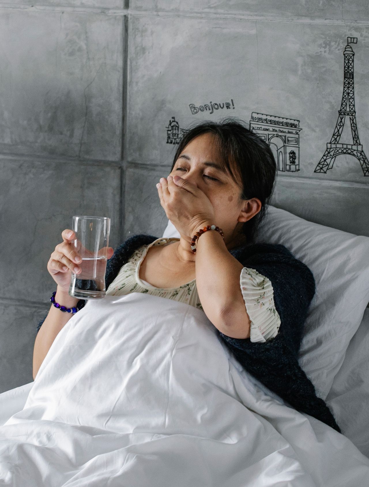 A woman lying in bed holds a glass of water while covering her mouth, appearing unwell.