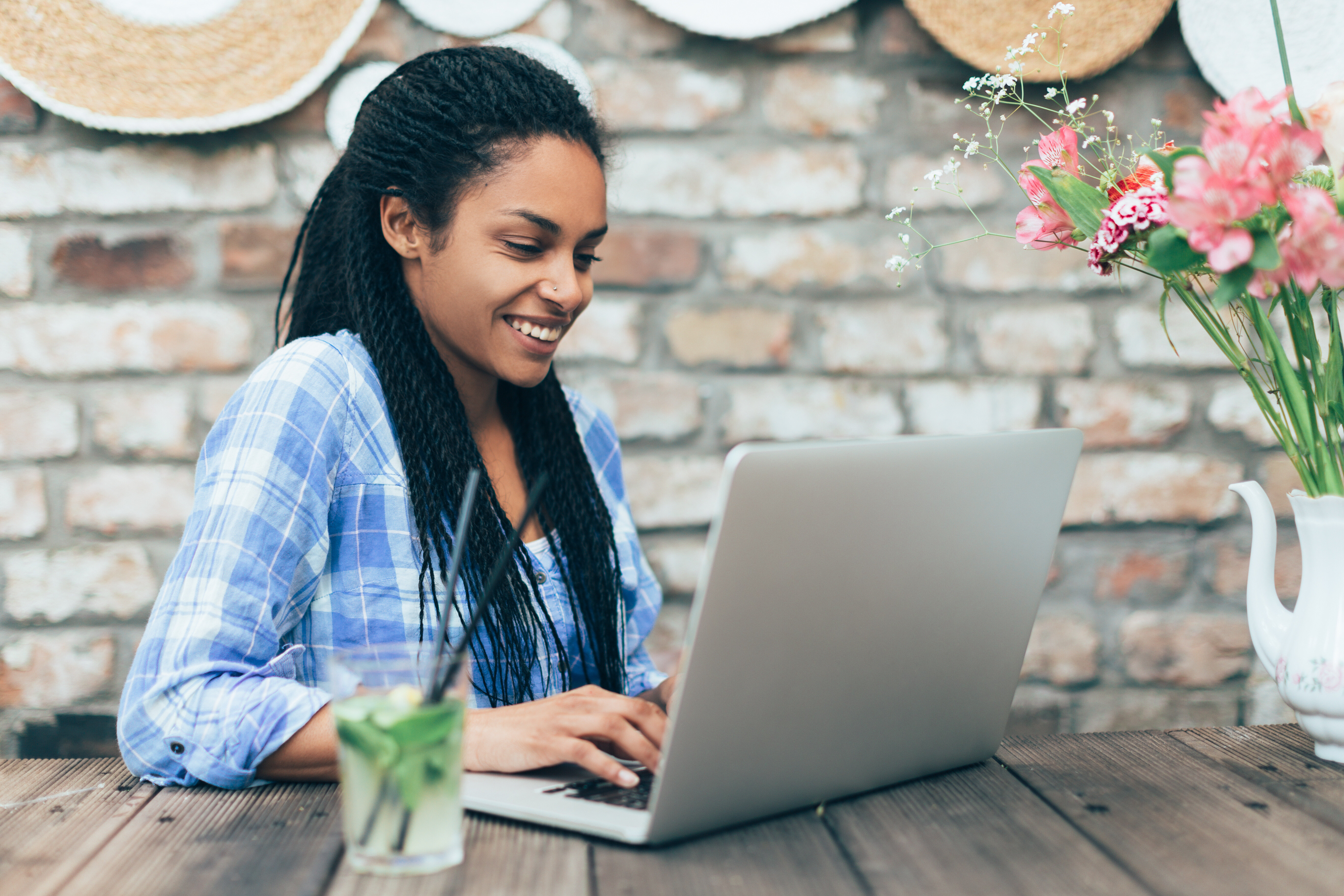 Smiling woman typing on a laptop at an outdoor wooden table with flowers and a brick wall backdrop