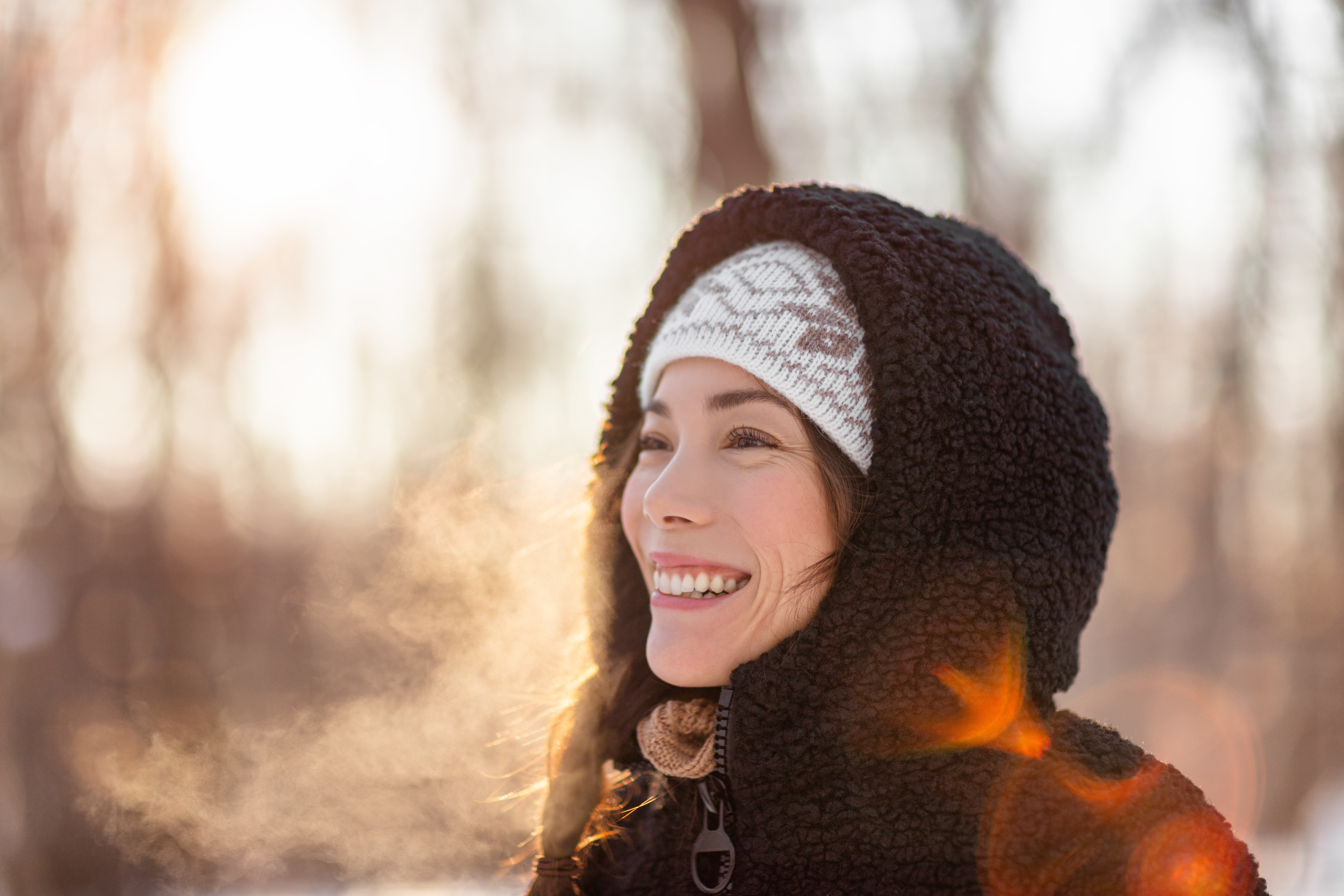 A smiling woman wearing a knit hat and scarf stands outdoors in winter light, looking up and to the side.