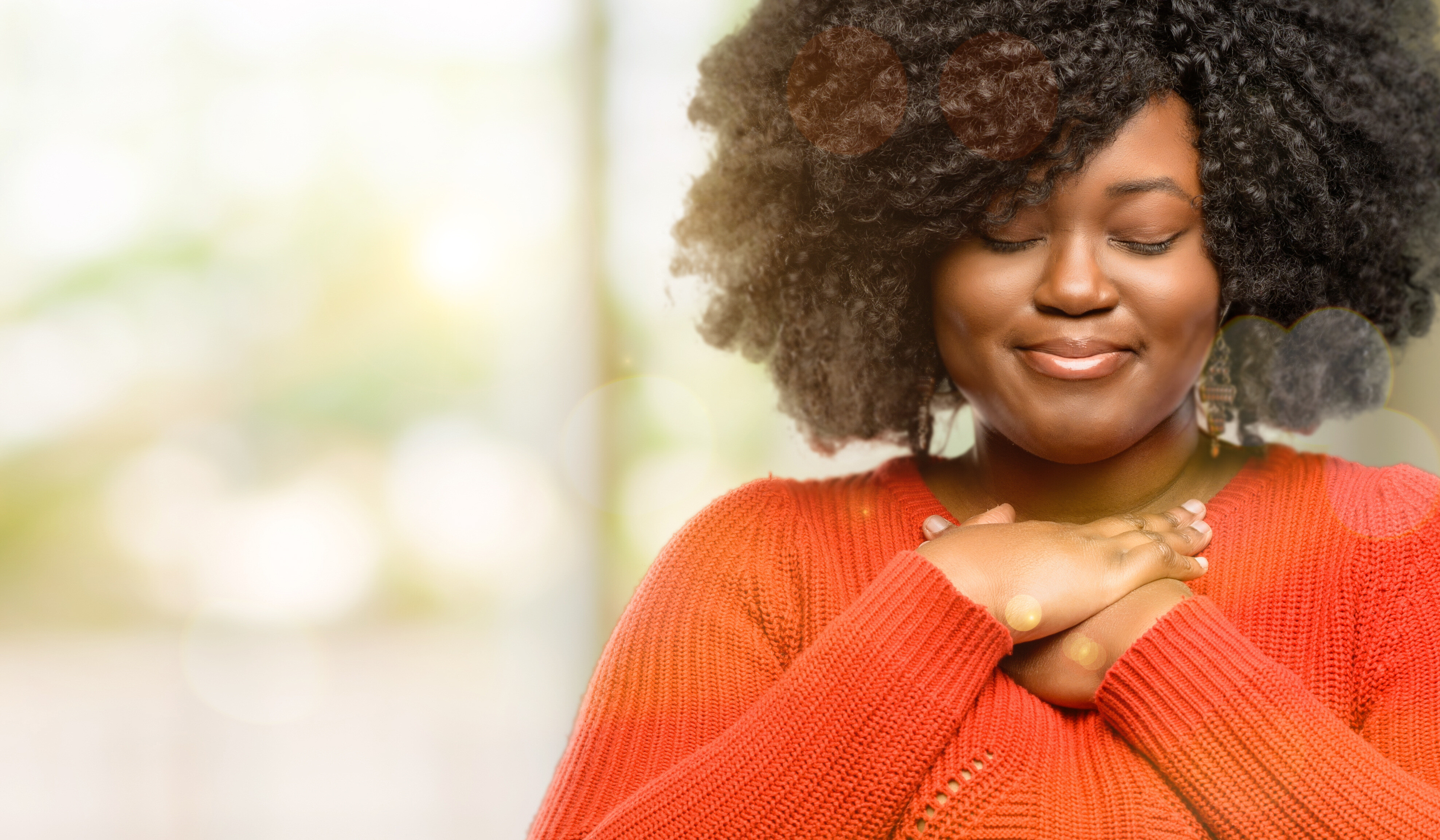 Woman with curly hair smiling with hands clasped over her heart, feeling grateful and at peace
