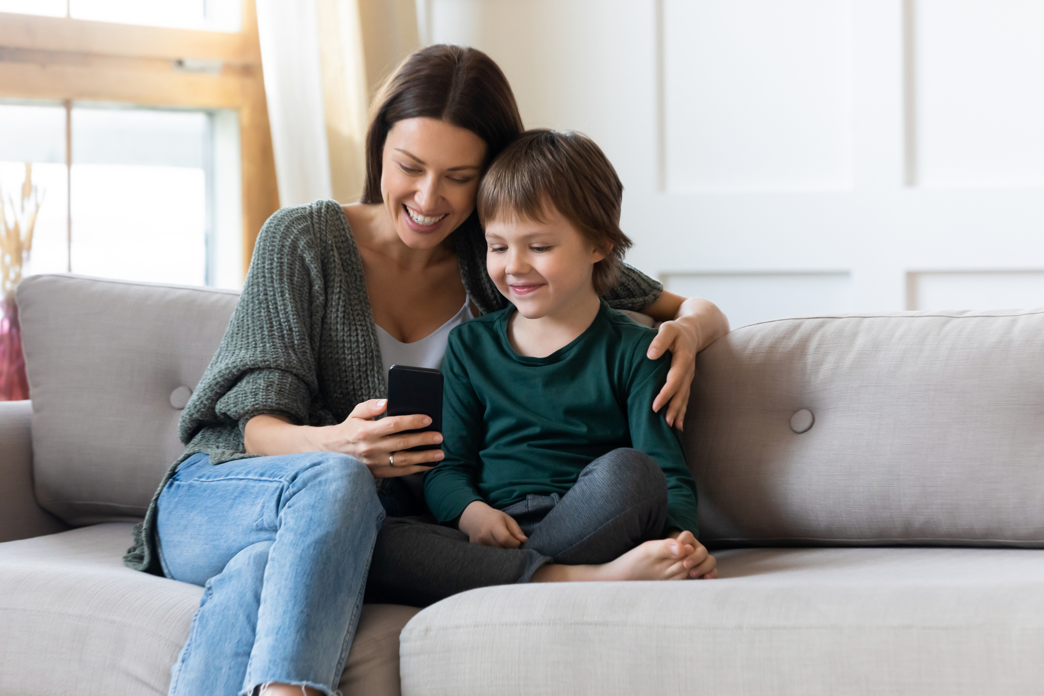 A smiling woman and a young child sit together on a couch, looking at a smartphone.