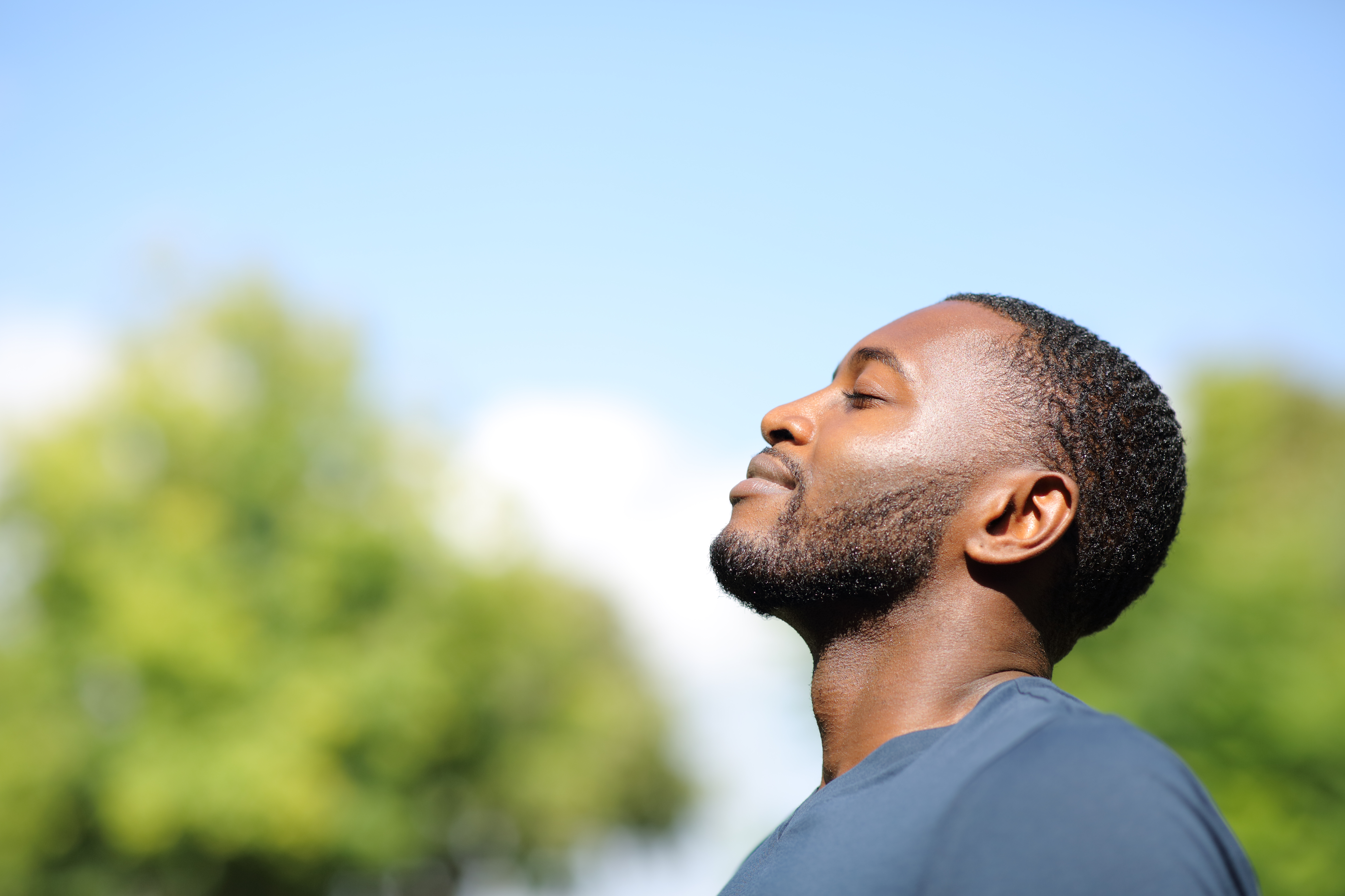 Man breathing fresh air outdoors with eyes closed, surrounded by greenery under a blue sky