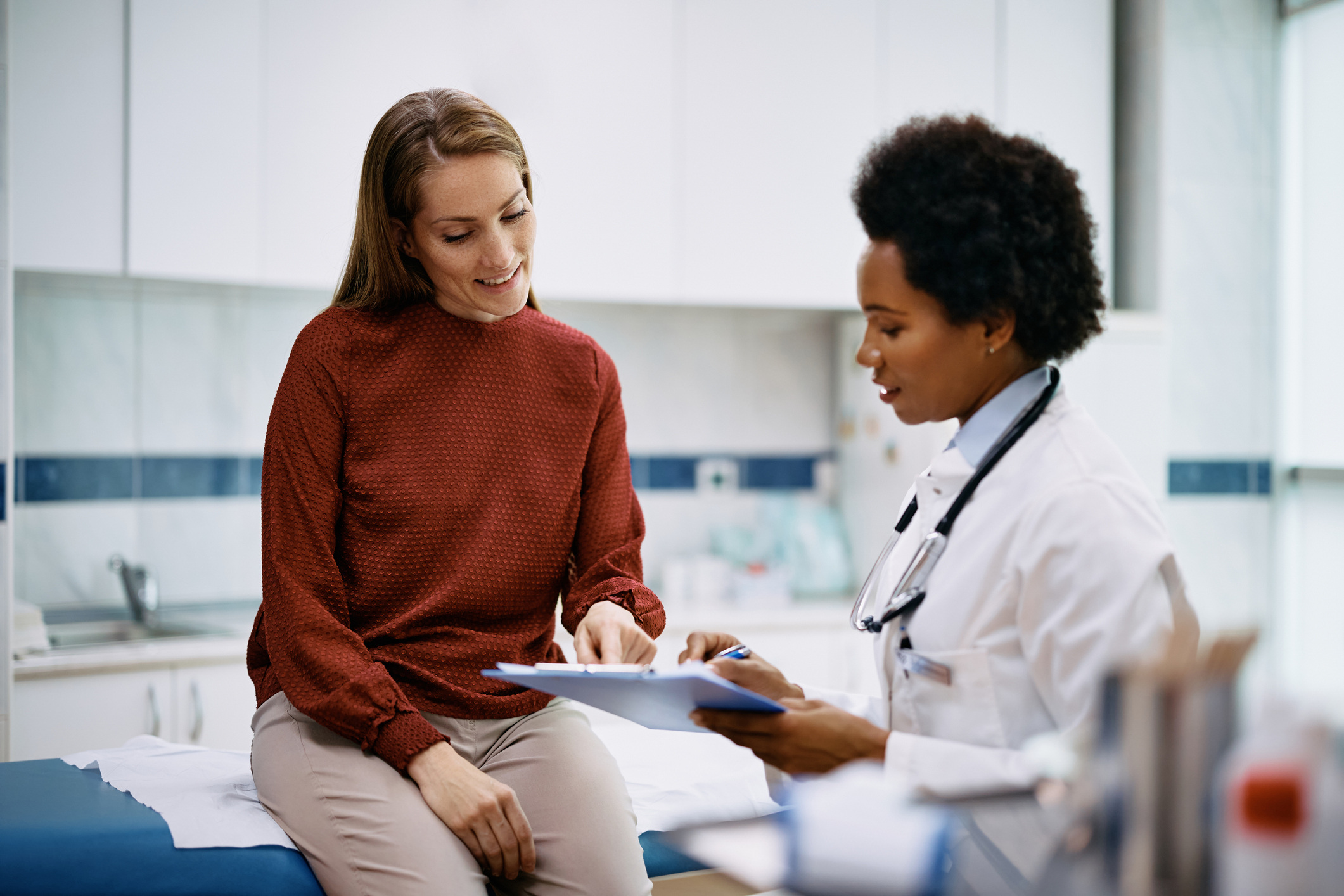 A patient sits on an exam table looking down while a doctor reviews notes on a clipboard during a consultation.