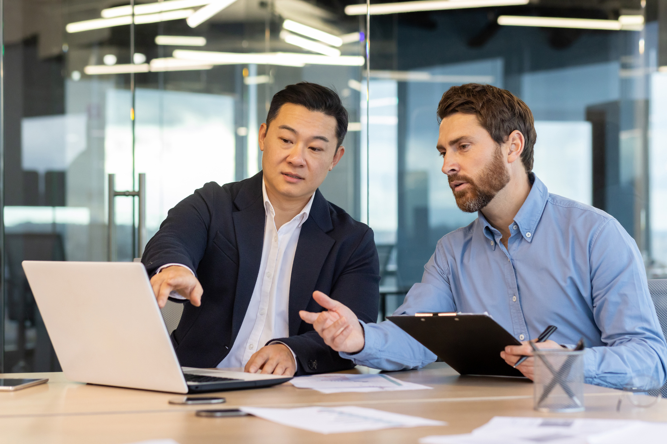 Two businessmen discussing work at a laptop in a modern glass-walled office, one holding a clipboard