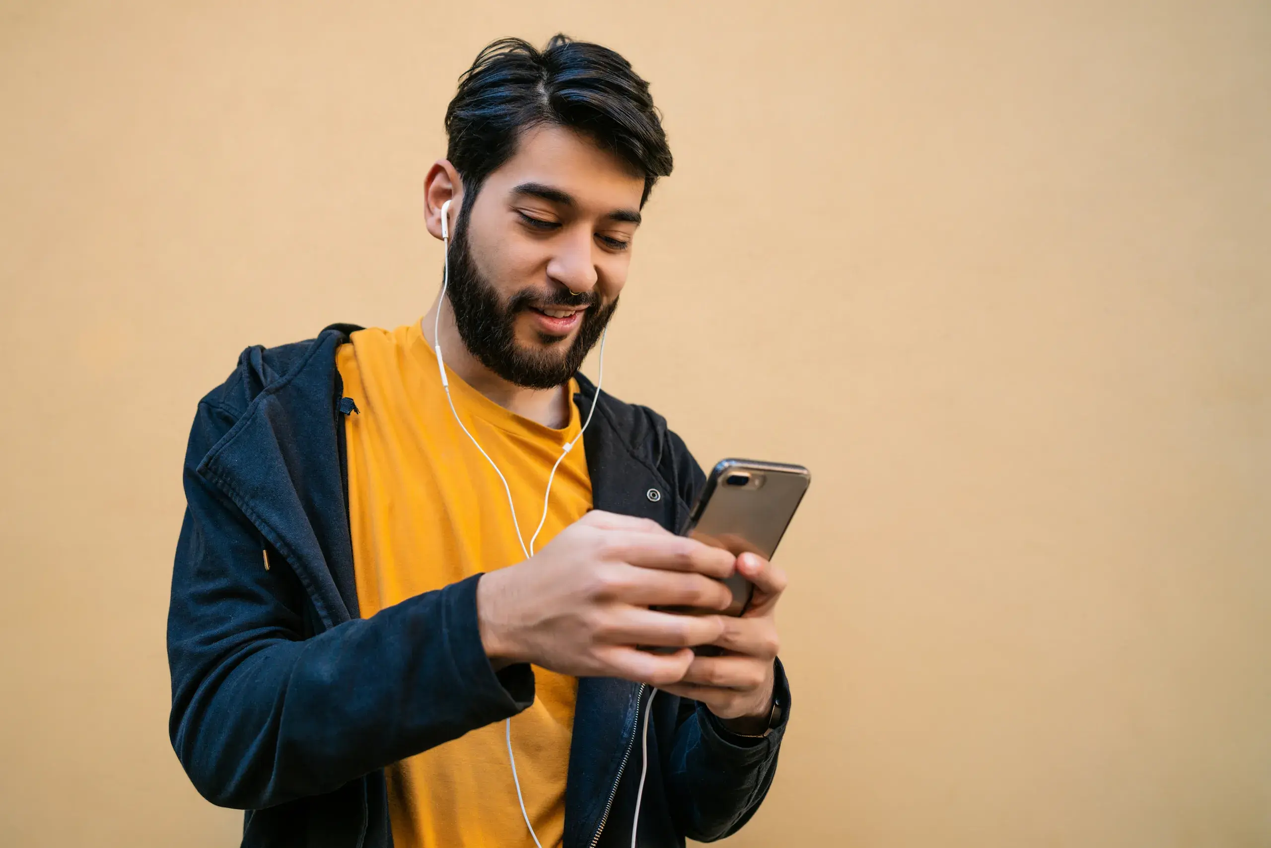 A bearded man wearing a mustard yellow top and dark jacket looks at his phone, standing against a tan background.
