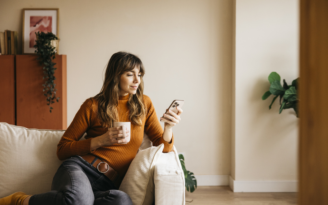 Woman sitting on a sofa at home holding a mug and checking her phone