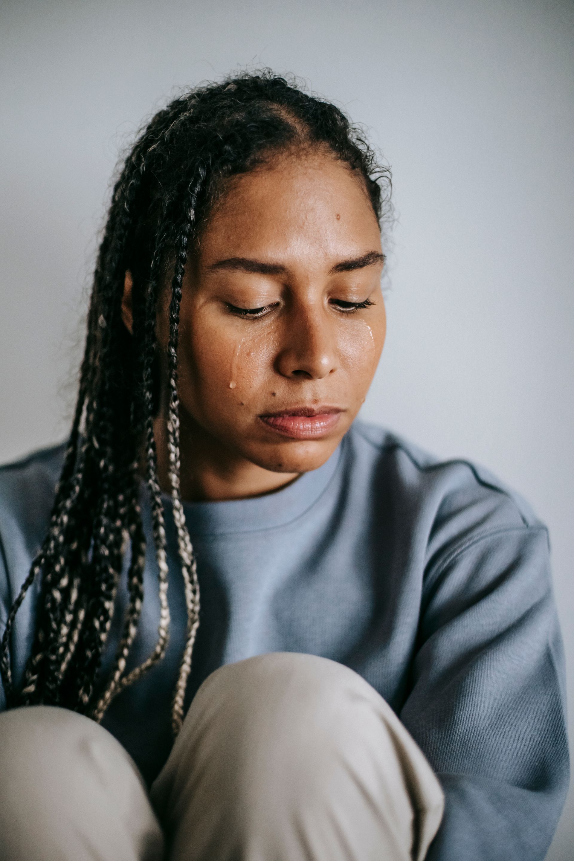 A woman with braided hair, wearing a grey sweatshirt, looks downward with a serious expression against a plain background.