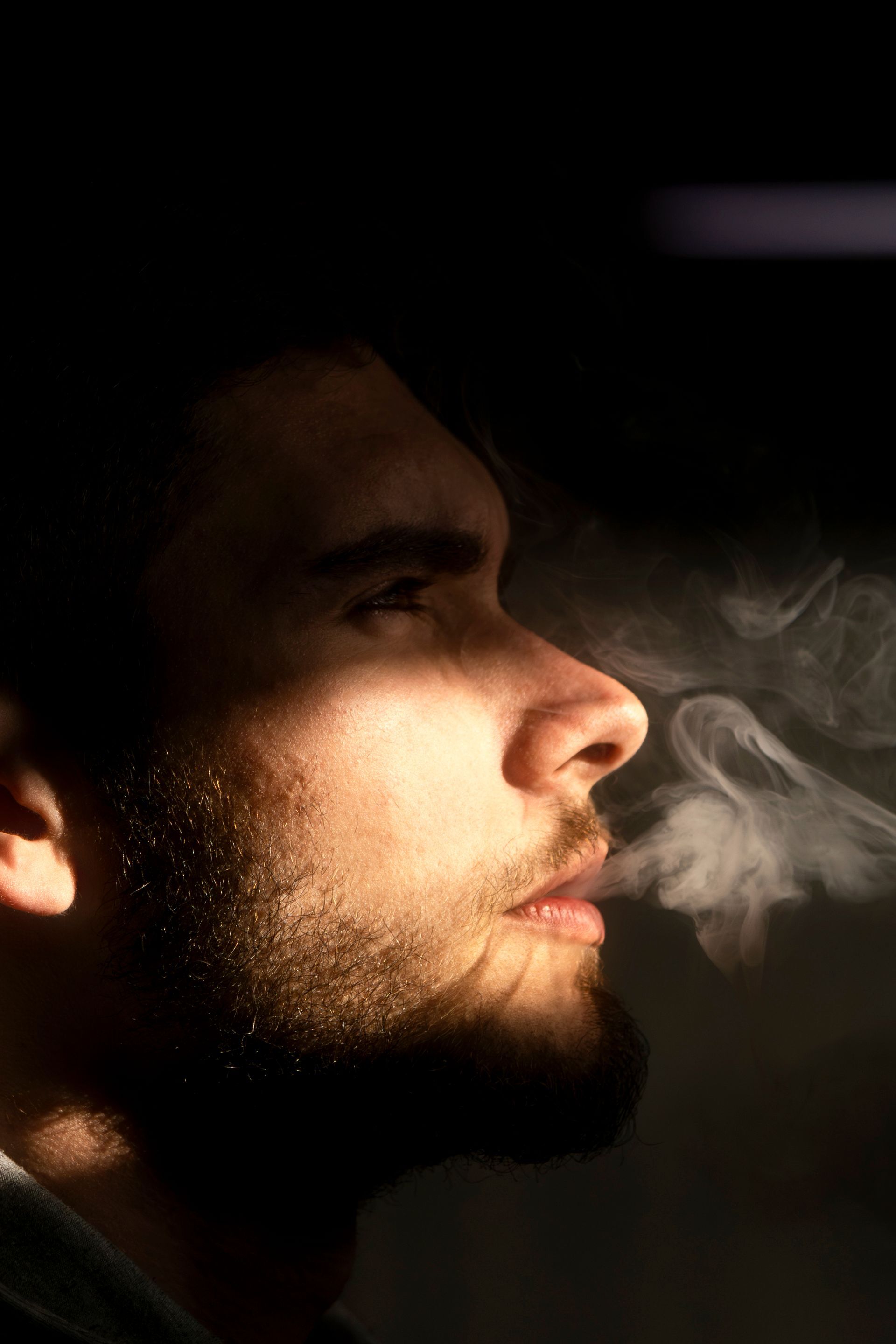 Profile close-up of a bearded man's face lit dramatically against a dark background, exhaling a visible cloud of smoke or vapor