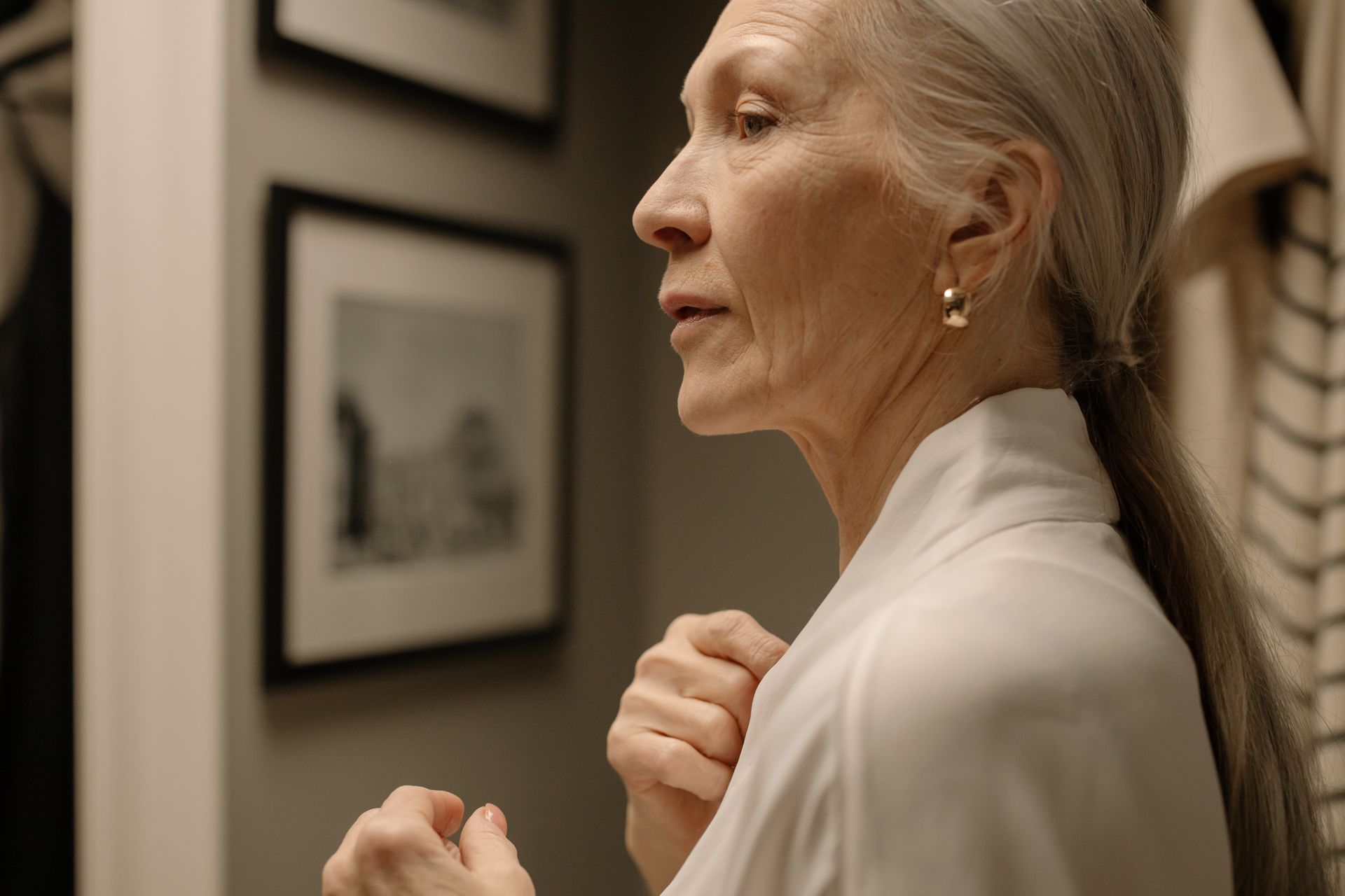 Profile view of an older woman with her hands raised near her chest, standing indoors near framed pictures on the wall. An orange 'Featured' badge is in the top right corner.