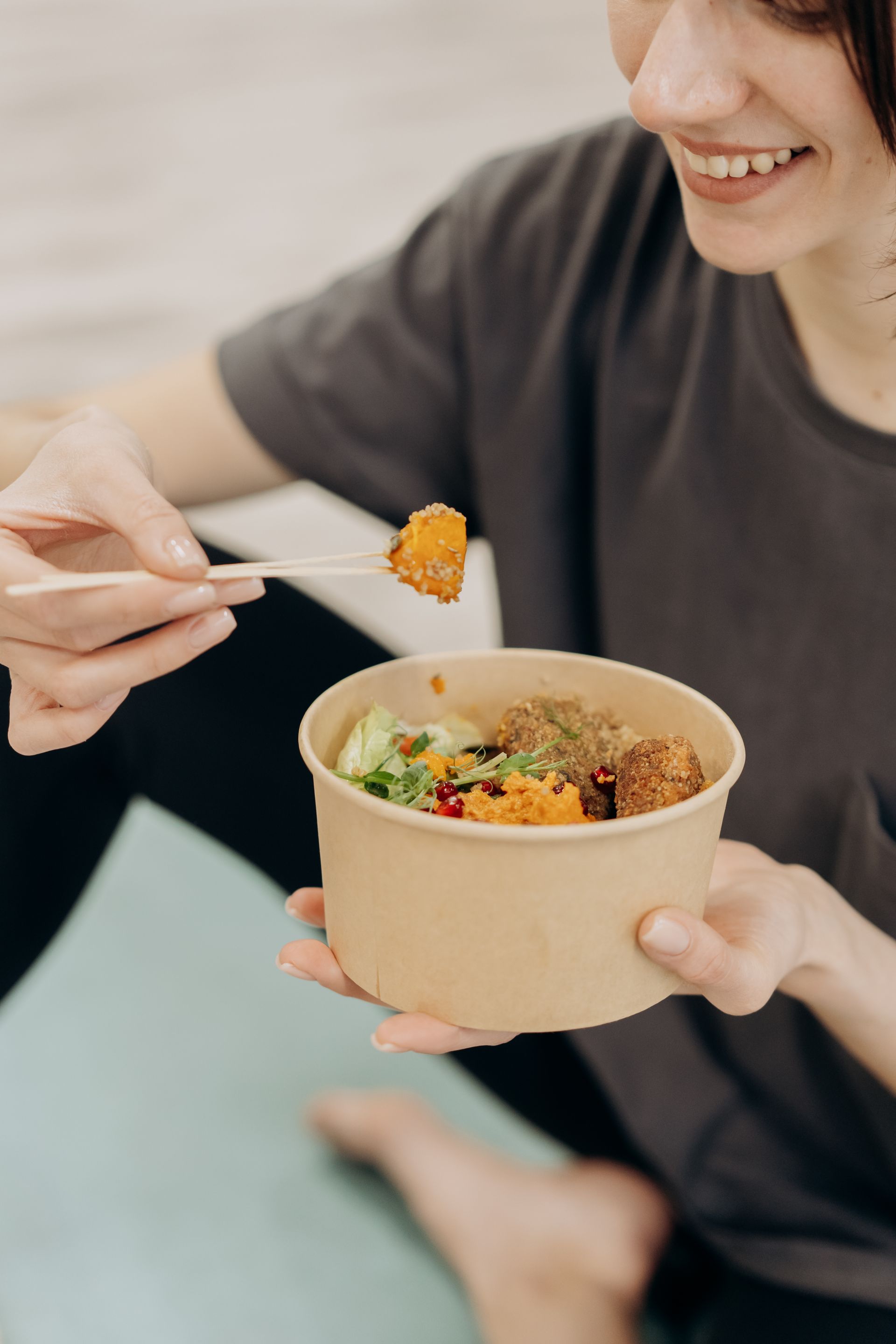 Close-up of hands holding a takeaway bowl of food with grains and vegetables, while another hand serves a piece of food into it with a fork.