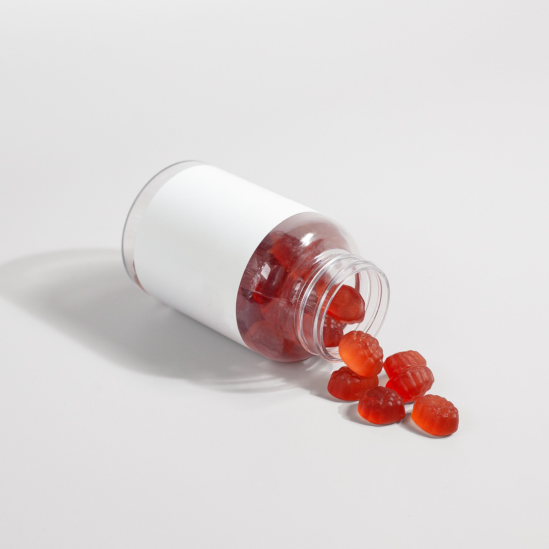 A tipped-over white pill bottle with orange-red softgel capsules spilling out onto a plain white surface.