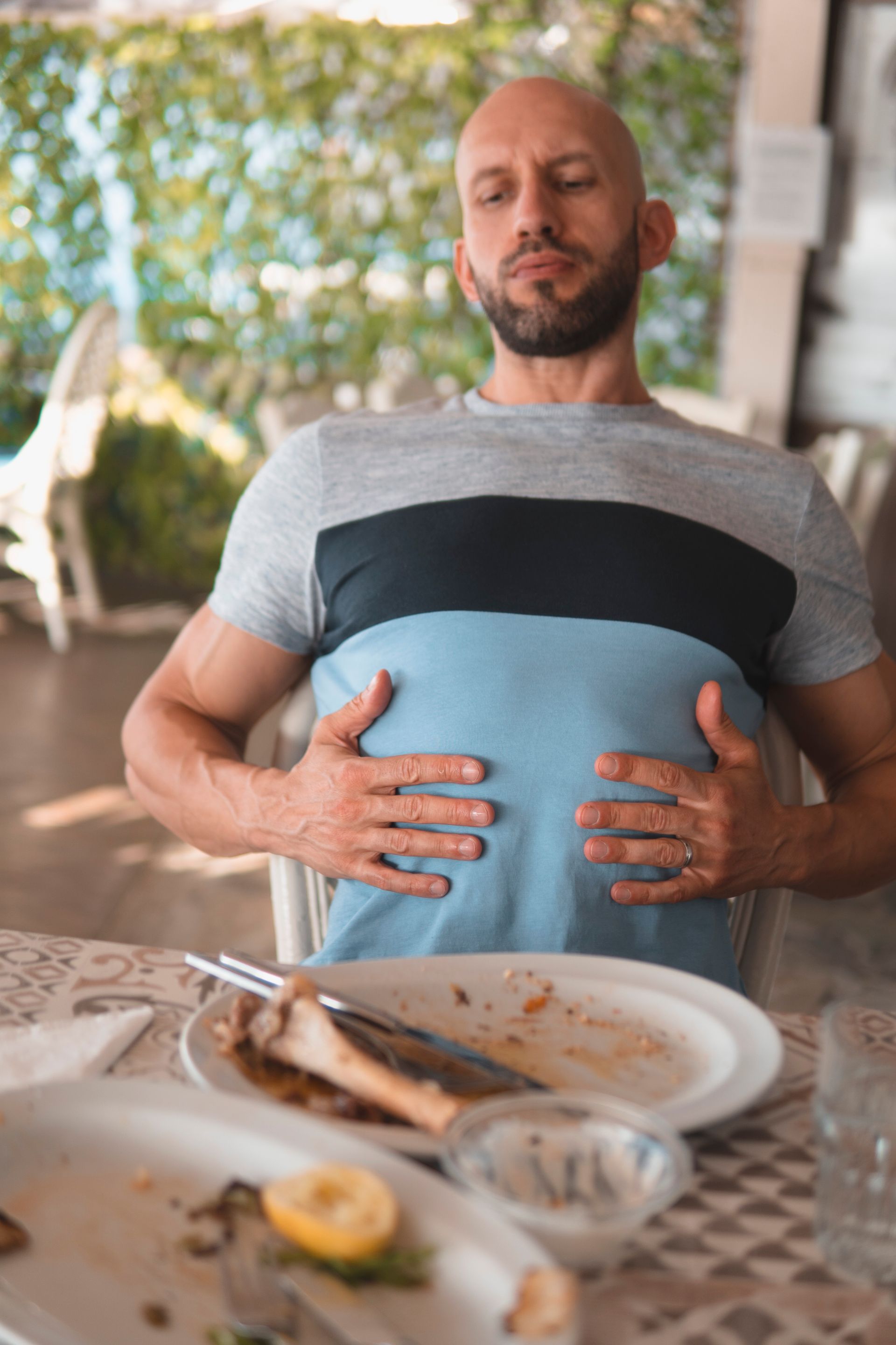 A man sitting at an outdoor dining table holds both hands on his stomach, suggesting fullness or discomfort after a meal, with a plate of food in front of him